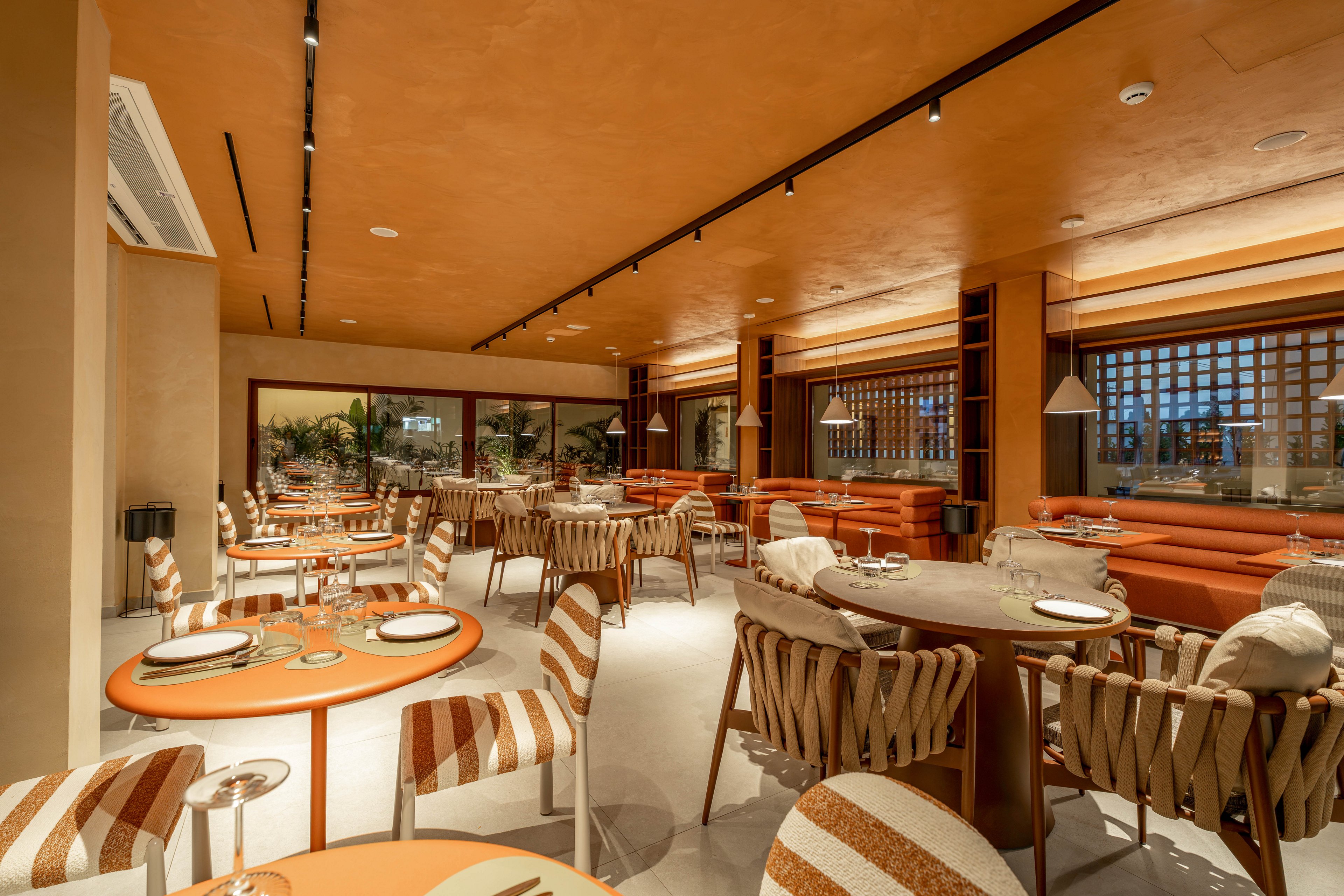 A modern restaurant interior features neatly arranged tables and chairs in a warm, earthy color palette. The space is decorated with striped upholstered chairs, round tables, and soft ambient lighting from pendant lamps. Large windows and glass partitions open the view to greenery outside, enhancing the inviting atmosphere. The overall design feels cozy yet sophisticated, ideal for relaxed dining.