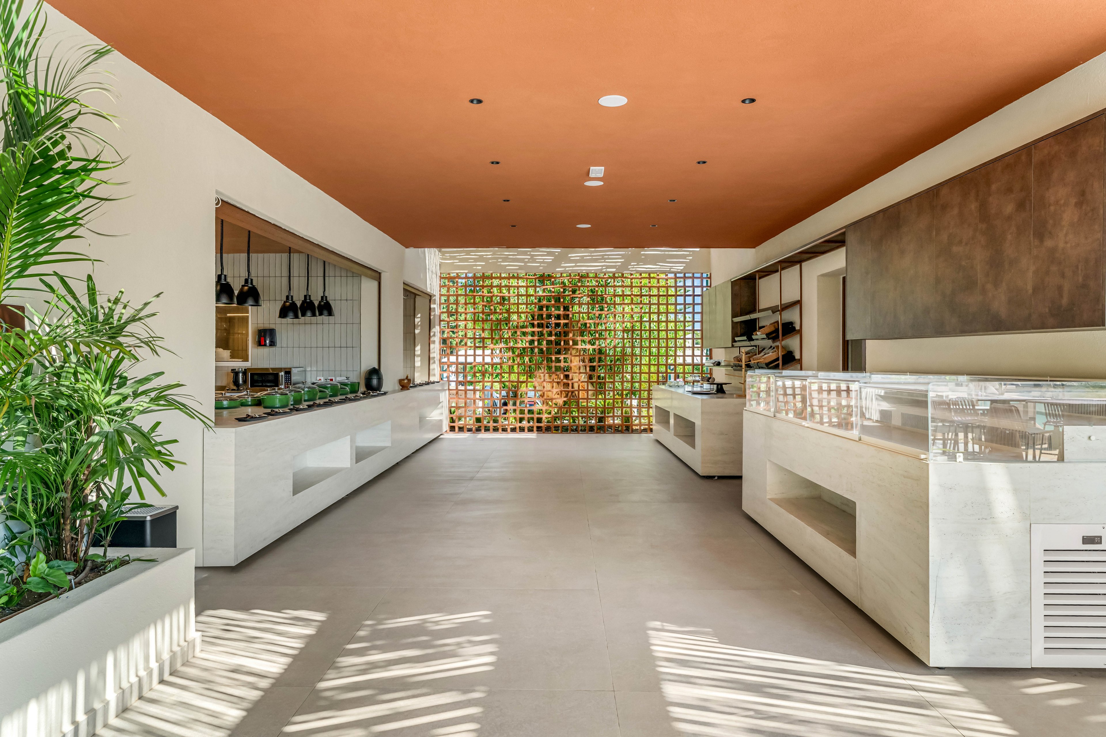 A contemporary buffet-style restaurant interior features long white counters and glass display cases on both sides of a wide aisle. Large potted plants and greenery add a fresh touch to the minimalist space. Warm terracotta ceiling tones contrast with light beige floors and walls. At the far end, a decorative lattice wall reveals palm trees and bright daylight outside, creating an airy, tropical atmosphere.