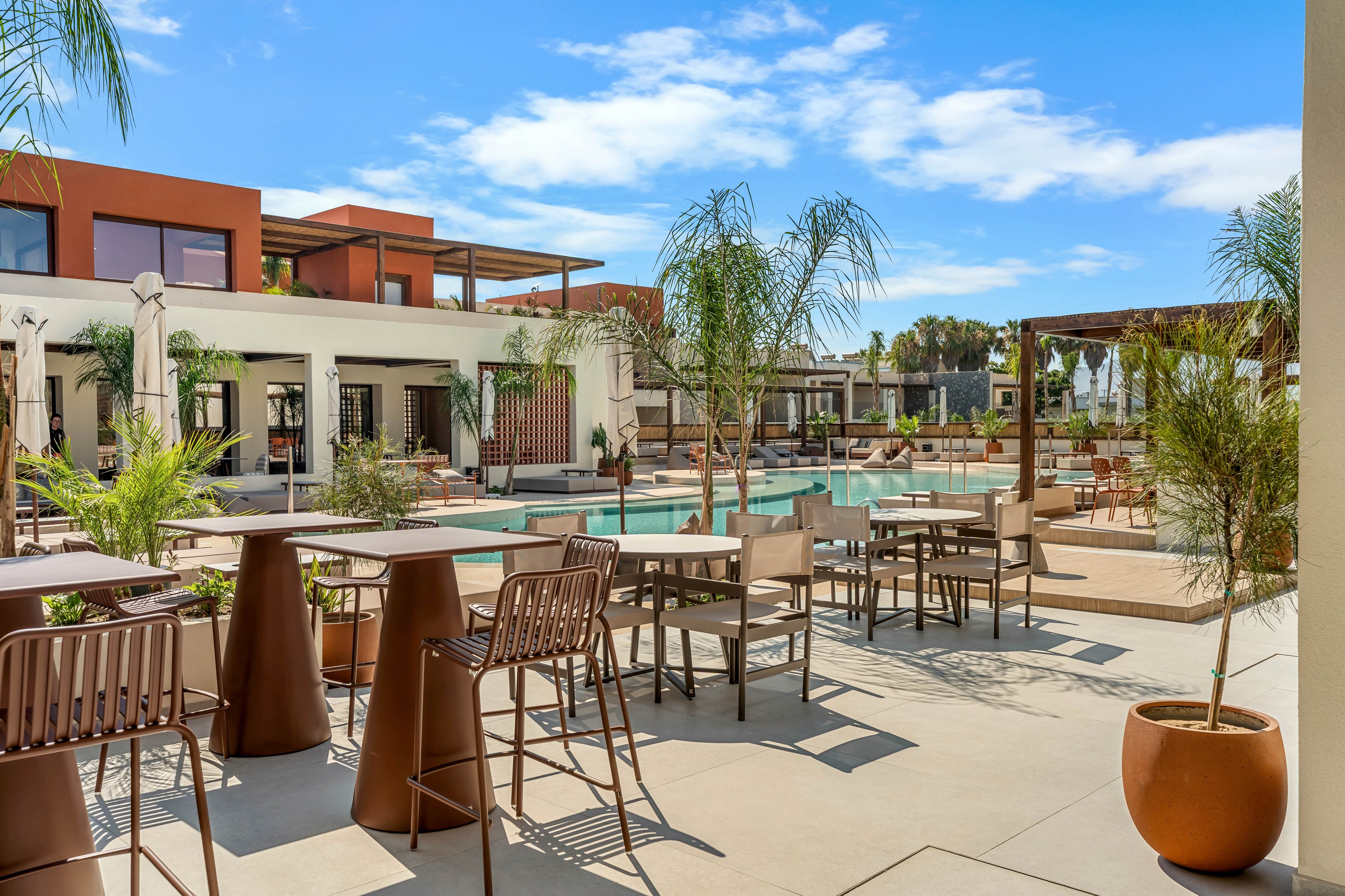 An outdoor resort-style patio features contemporary tables and chairs arranged around a swimming pool. The setting includes palm trees, potted plants, and shaded pergolas under a bright blue sky with scattered clouds. Warm earth tones and clean architectural lines create a modern, inviting atmosphere. The scene appears calm and unoccupied, emphasizing relaxation and leisure by the pool.