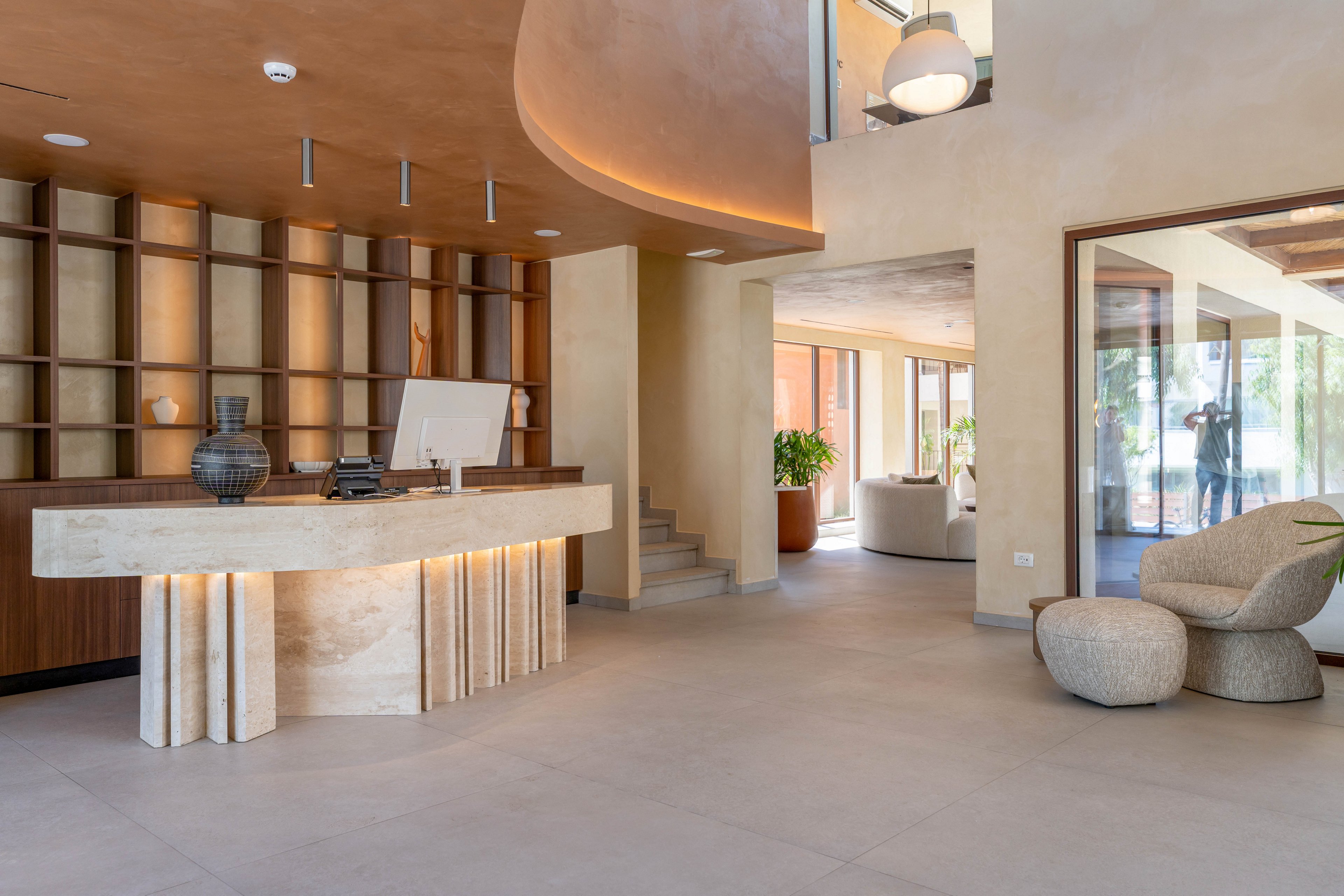 A contemporary reception desk stands in a spacious, minimalist lobby. The area features warm neutral tones, smooth stone surfaces, and built-in wooden shelving behind the counter. Soft indirect lighting highlights the curved architectural details and open layout. In the background, lounge seating, indoor plants, and large windows create an inviting, airy atmosphere.