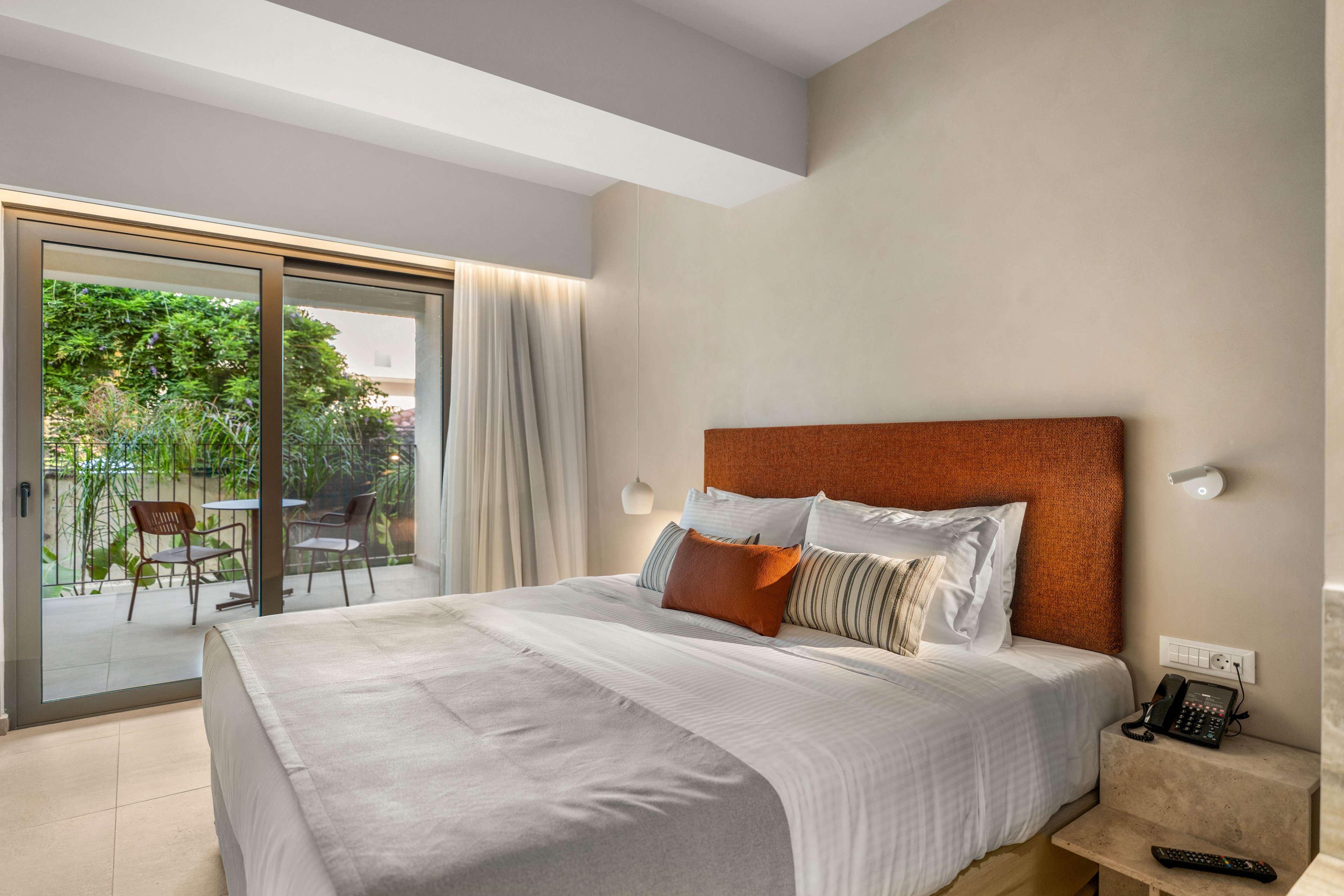 A modern hotel-style bedroom features a large neatly made bed with white linens and accent pillows against an upholstered headboard. The room opens onto a small terrace through sliding glass doors, revealing greenery and an outdoor seating area. Soft neutral walls, minimal decor, and warm lighting create a calm, inviting atmosphere. A bedside table with a telephone and wall-mounted reading lights complete the contemporary interior.