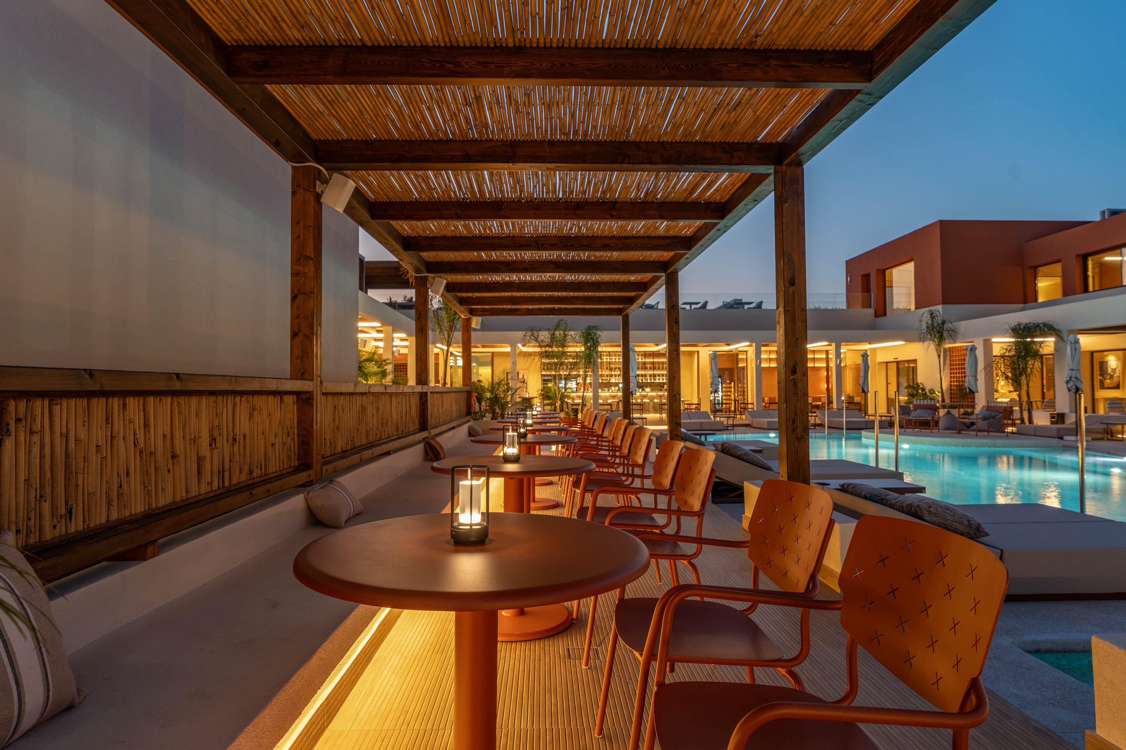 An elegant outdoor lounge bar overlooks a tranquil swimming pool at dusk. Warm ambient lighting glows under the bar counter and from small lanterns placed on round tables. Orange chairs line the bar beneath a wooden pergola with a woven roof. In the background, modern resort buildings and palm trees are softly illuminated, reflecting in the calm pool water.
