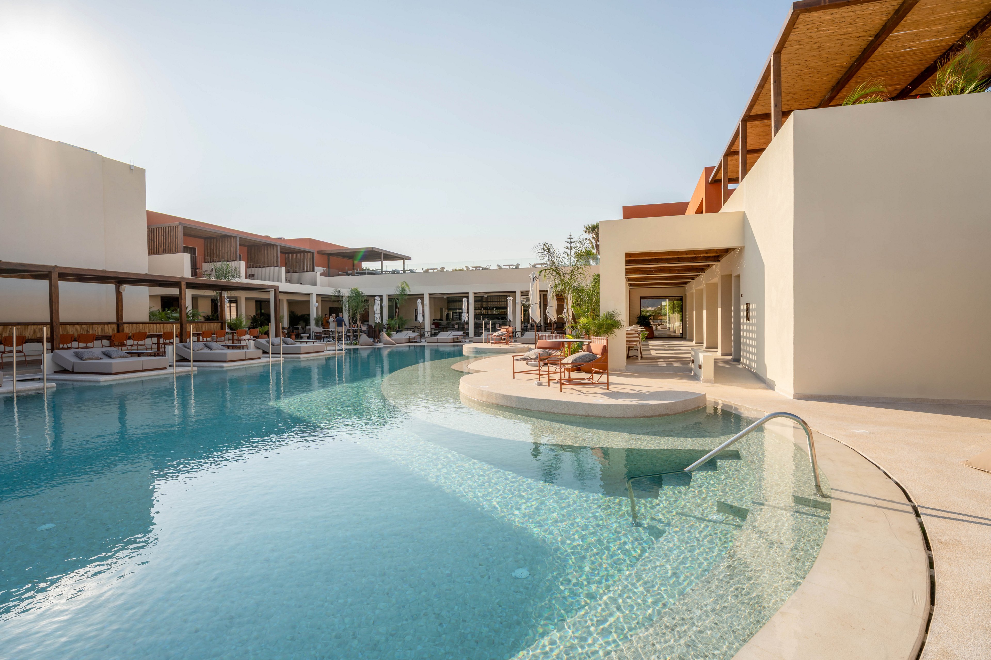 A large, curved swimming pool sits at the center of a contemporary resort complex. Minimalist cream-colored buildings with wooden pergolas and shaded seating areas surround the water. Sun loungers and umbrellas line the poolside, creating a relaxed vacation atmosphere under clear blue skies. The scene suggests a tranquil, luxurious getaway in a warm climate.