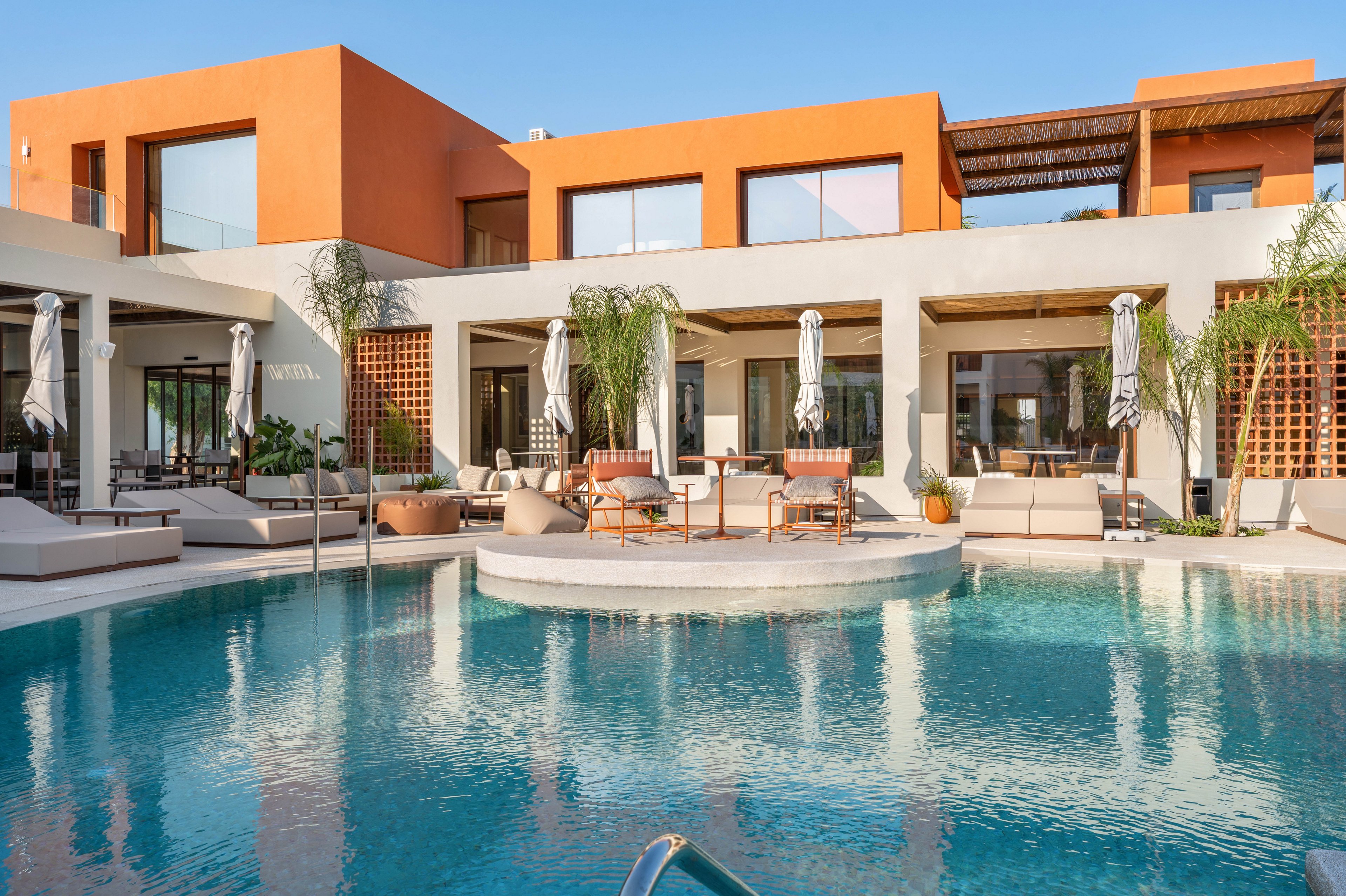 A contemporary villa with clean geometric lines overlooks a clear blue swimming pool. The building features white and terracotta-colored facades, large windows, and shaded terraces. Poolside lounge chairs, umbrellas, and palm trees create a relaxed resort atmosphere. The scene is captured in bright daylight under a clear blue sky, emphasizing the modern, luxurious setting.