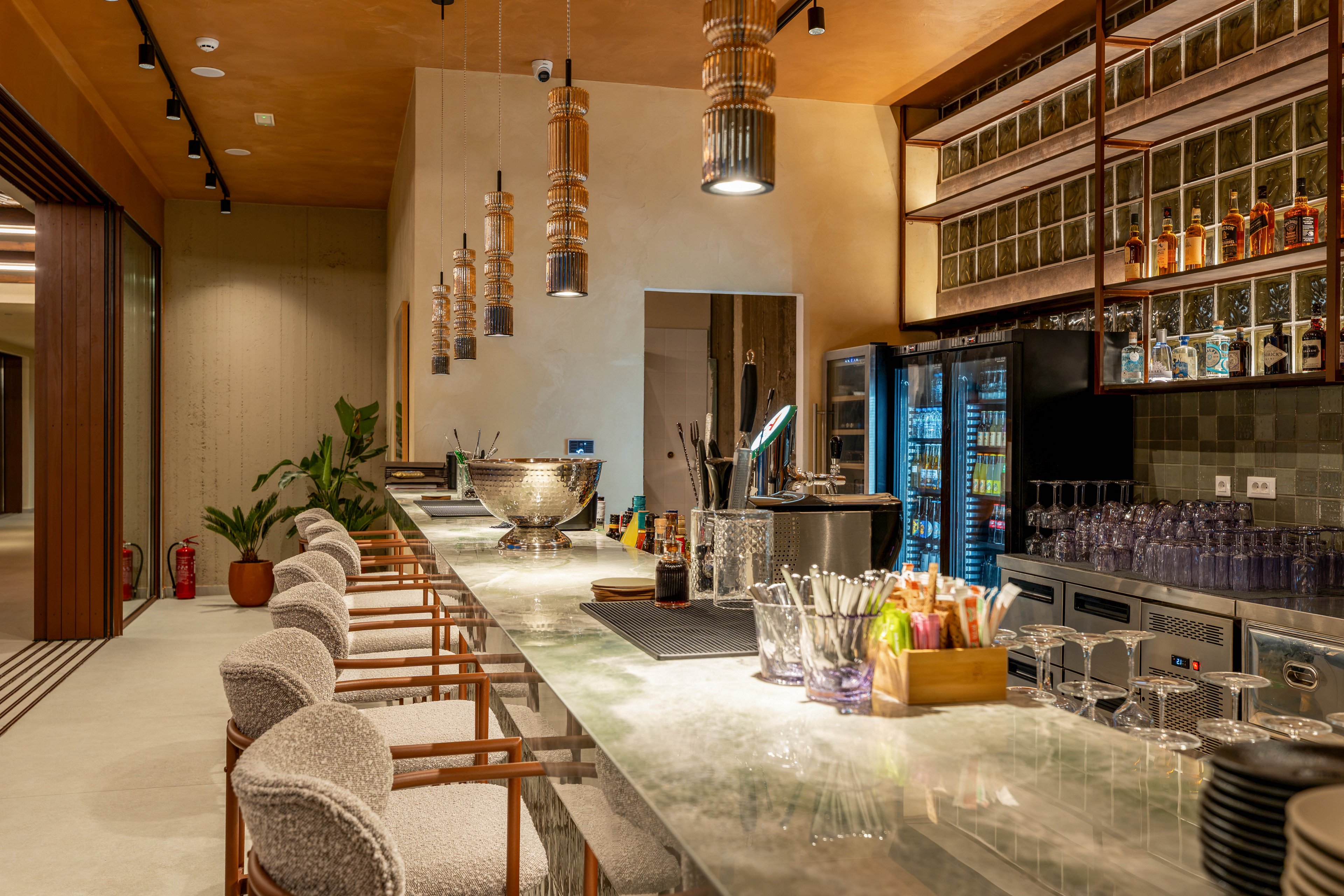 A contemporary bar interior features a long marble counter lined with cushioned barstools. The setting includes hanging pendant lights, glass shelving with bottles, and refrigerated drink displays. Bar tools, glassware, and garnishes are neatly arranged on the countertop, suggesting a ready-to-serve cocktail environment. Warm lighting and neutral tones create an inviting, upscale atmosphere in this modern hospitality space.