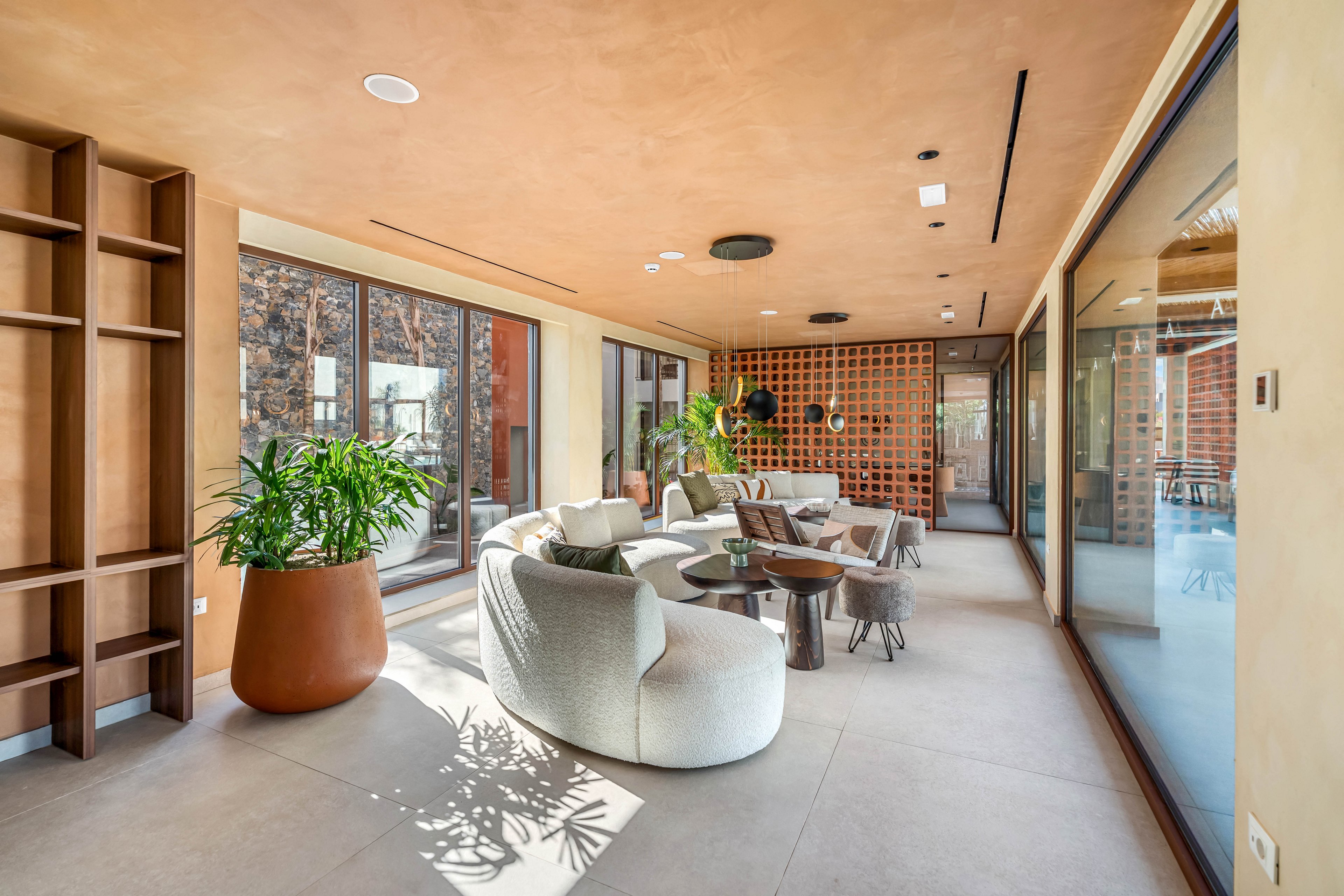 A bright modern lounge features curved upholstered chairs and a low round coffee table arranged for conversation. Floor-to-ceiling glass doors open to an outdoor area, filling the room with natural light and reflections. Warm earth-toned walls and ceiling contrast with light stone flooring and a decorative grid partition. Large potted plants add greenery and soften the clean architectural lines. The space feels airy, inviting, and designed for relaxed social gatherings.