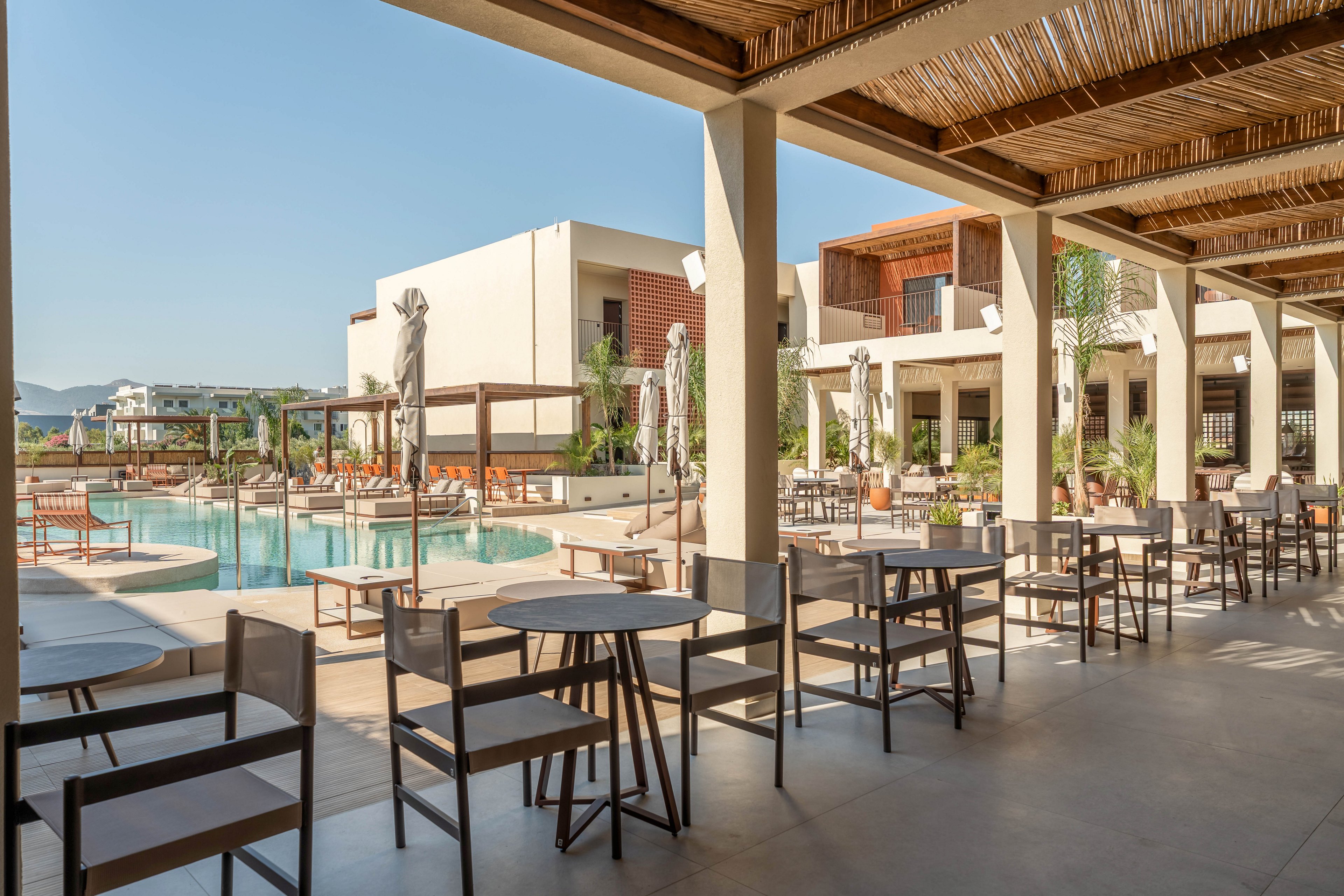 A contemporary resort terrace overlooks a clear swimming pool surrounded by sun loungers and umbrellas. Simple wooden tables and chairs are neatly arranged under a shaded pergola, creating an inviting outdoor dining space. The modern buildings in the background feature clean lines, balconies, and large windows. Bright daylight and blue skies enhance the relaxed, vacation atmosphere of the scene.