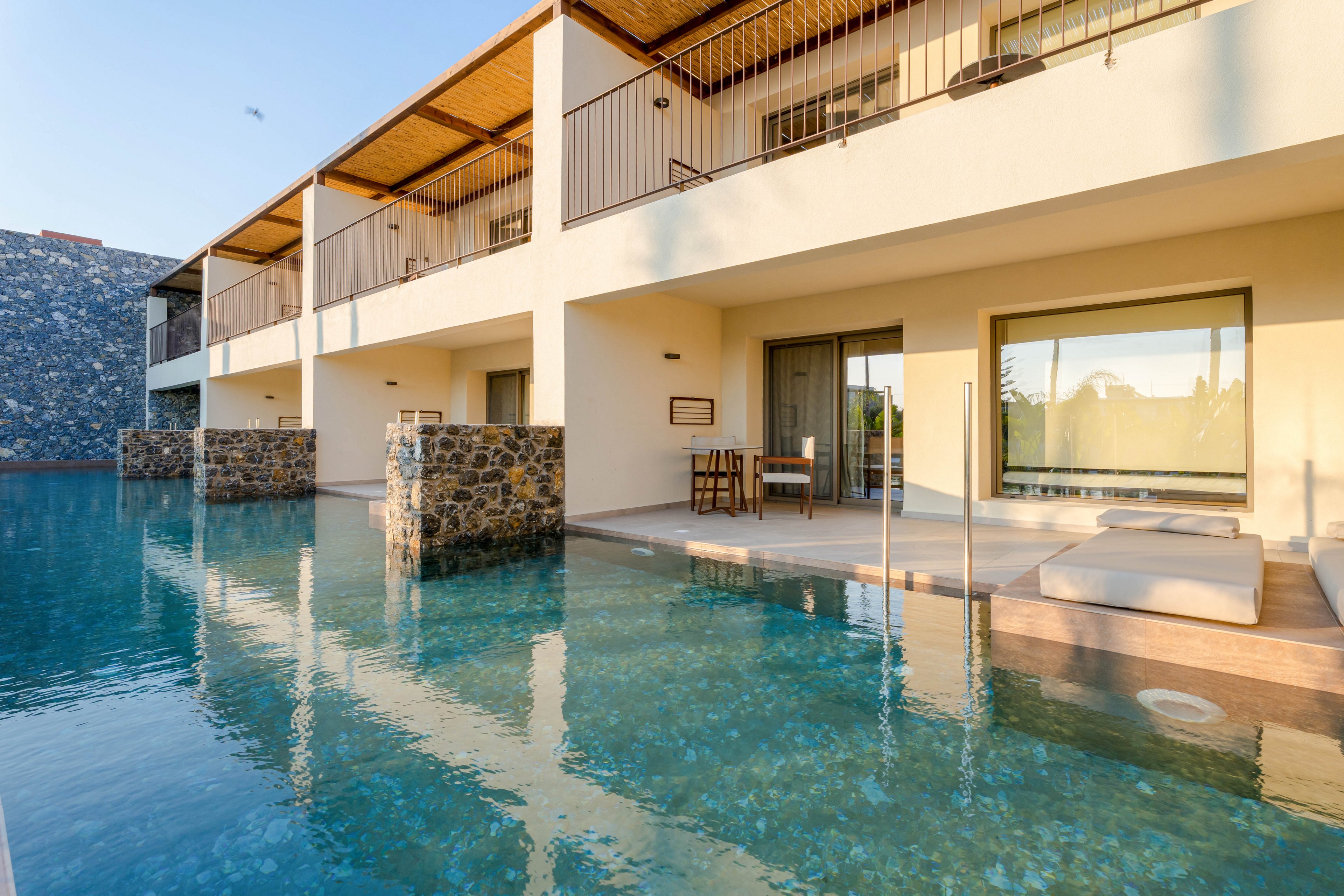 A contemporary hotel building features ground-floor rooms opening directly onto a tranquil swimming pool. Private terraces with lounge chairs and a small table create an inviting outdoor living space. The clear blue water reflects the warm tones of the building and the wooden ceiling above the balconies. Soft evening light enhances the calm, resort-like atmosphere of the scene.