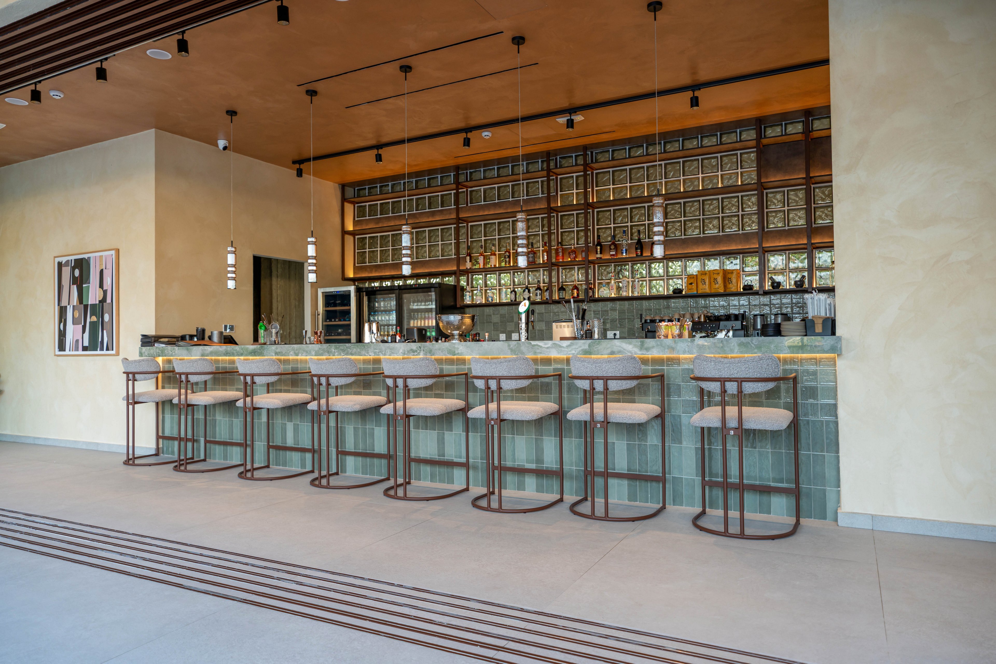 A contemporary bar interior features a long counter lined with cushioned high stools. The setting is clean and minimalist, with light-colored walls, a wooden ceiling, and pendant lights hanging above the bar. Glass shelving and neatly arranged bottles create a structured backdrop behind the counter. A framed abstract artwork decorates the wall, adding a subtle artistic touch to the calm, modern atmosphere.
