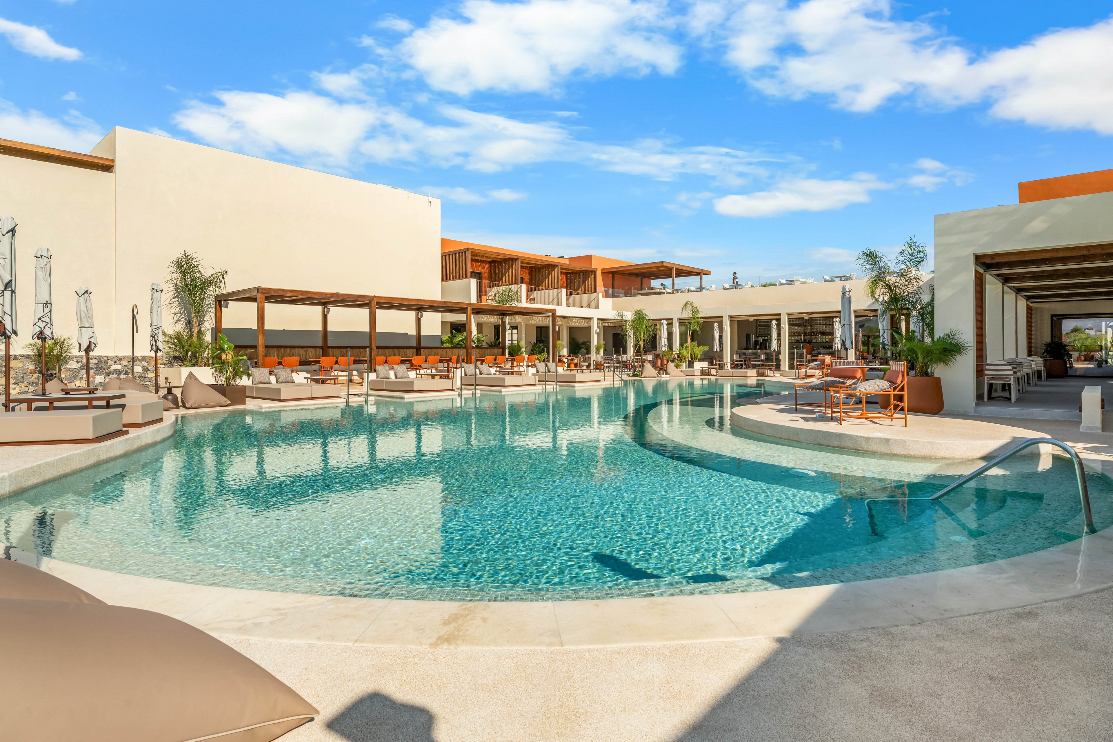A large, curved swimming pool sits at the center of a modern resort complex. Lounge chairs and shaded pergolas line the poolside, creating a comfortable relaxation area. Contemporary low-rise buildings with clean architectural lines surround the water. The bright blue sky and scattered clouds enhance the sunny, vacation-like atmosphere.