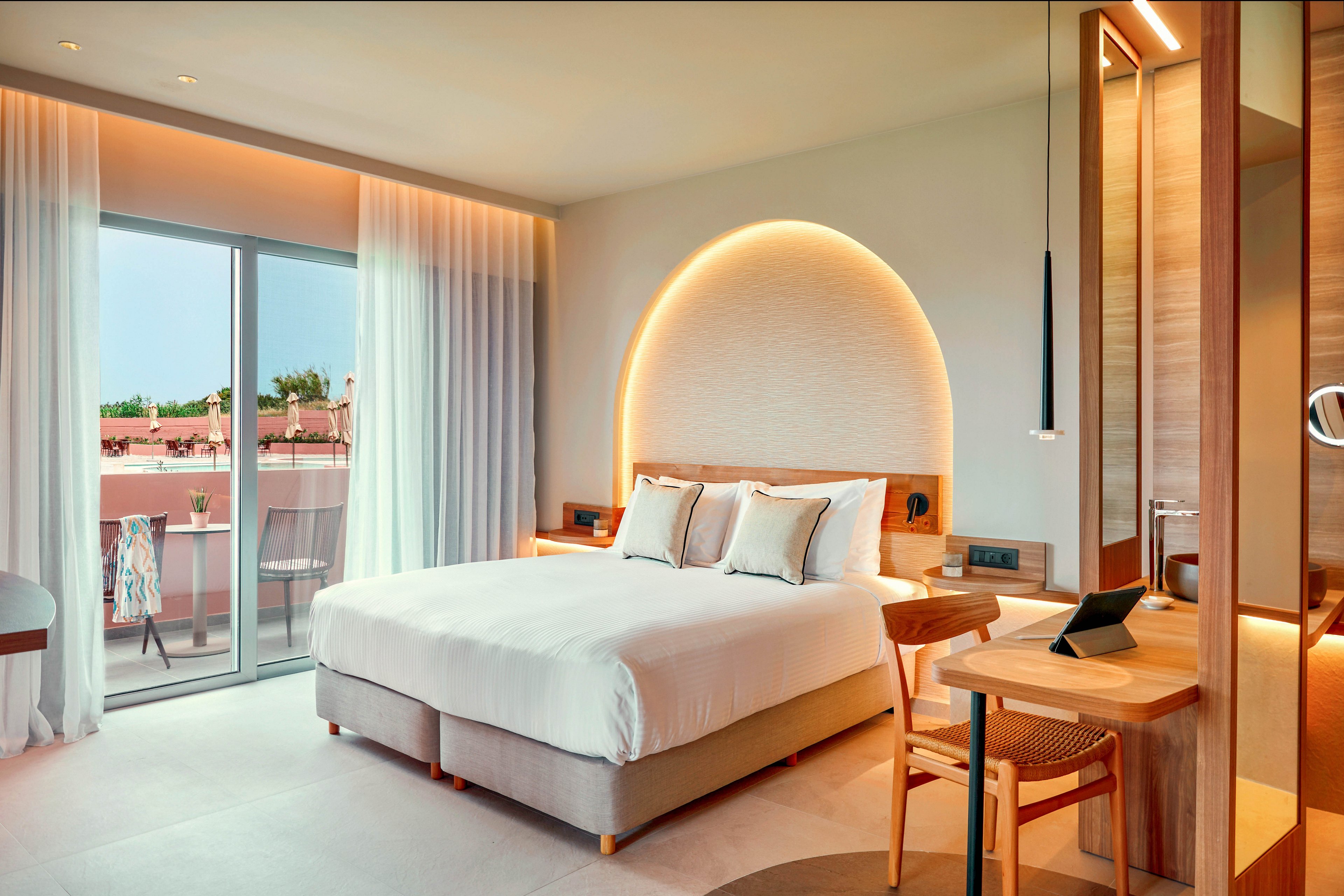 Contemporary hotel room featuring a large bed with white linens and accent pillows is shown in warm natural light. A sliding glass door with sheer curtains opens to a balcony overlooking pastel-colored buildings. The interior includes a wooden desk with chair, pendant light, and soft indirect lighting in an arched wall niche behind the bed. Overall, the space feels bright, minimalist, and inviting, ideal for a relaxing stay.