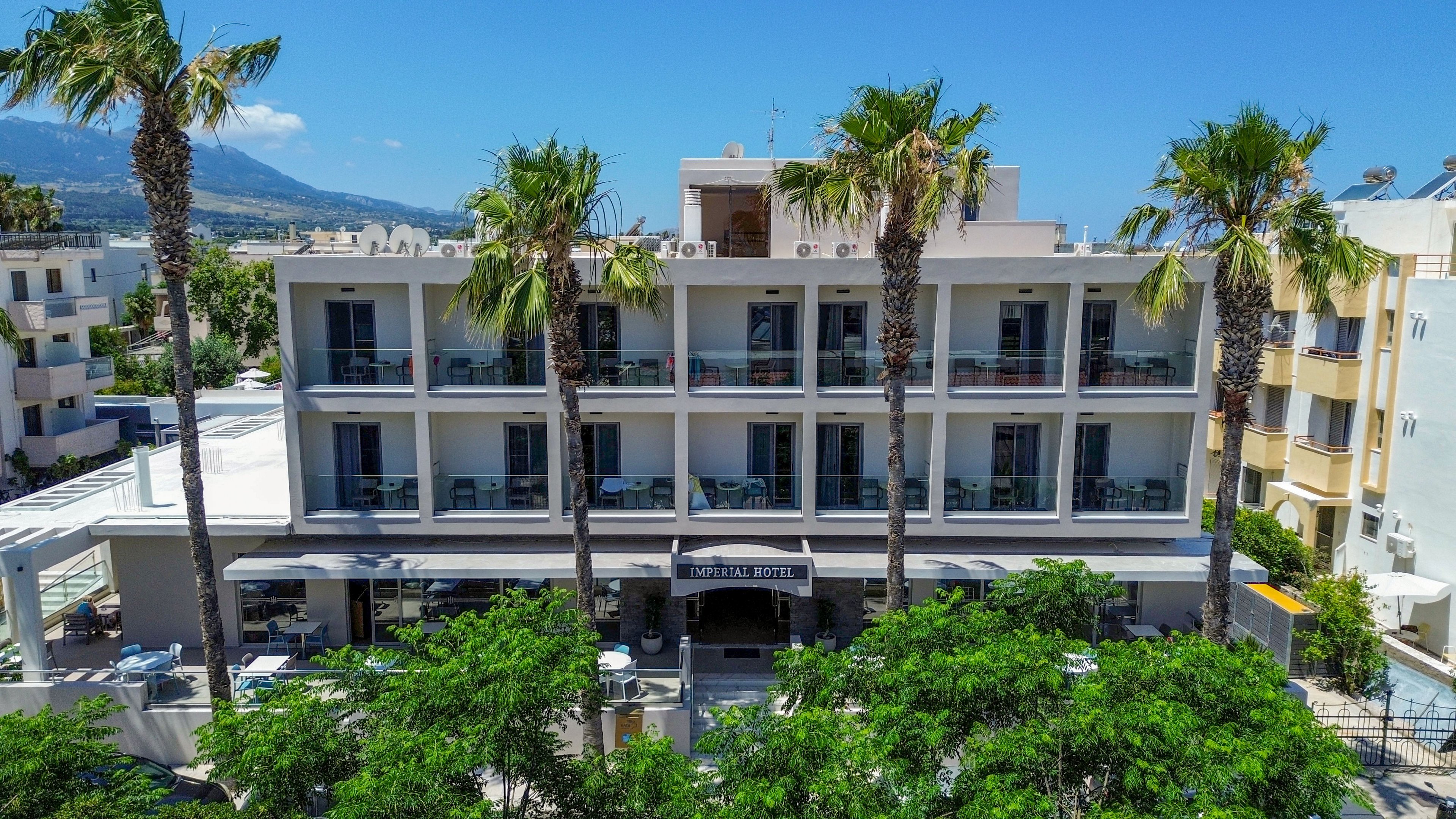 A contemporary multi-story hotel building is shown from the front, framed by tall palm trees and lush greenery. The hotel features balconies on each floor, with outdoor seating areas visible along the facade. Surrounding structures and a distant mountain range suggest a coastal or resort town setting under a clear blue sky. The scene conveys a relaxed vacation atmosphere with modern urban resort style.