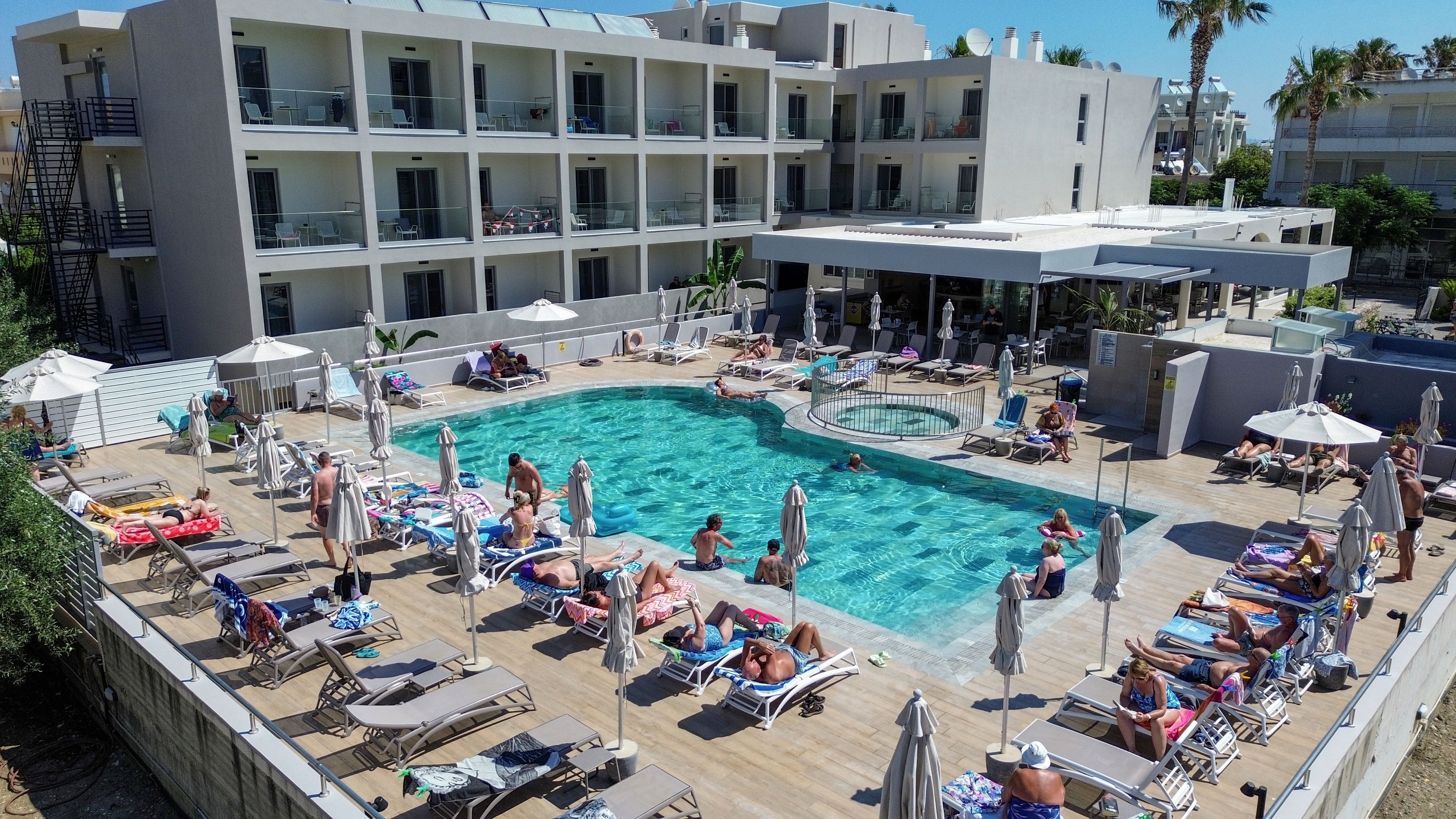 A large outdoor swimming pool is surrounded by numerous sun loungers and umbrellas at a modern hotel. Guests relax, sunbathe, and swim on a bright, clear day, creating a lively vacation atmosphere. The multi-story building with balconies overlooks the pool deck, emphasizing resort-style accommodation. Palm trees and contemporary architecture frame the scene, suggesting a warm coastal or holiday destination.