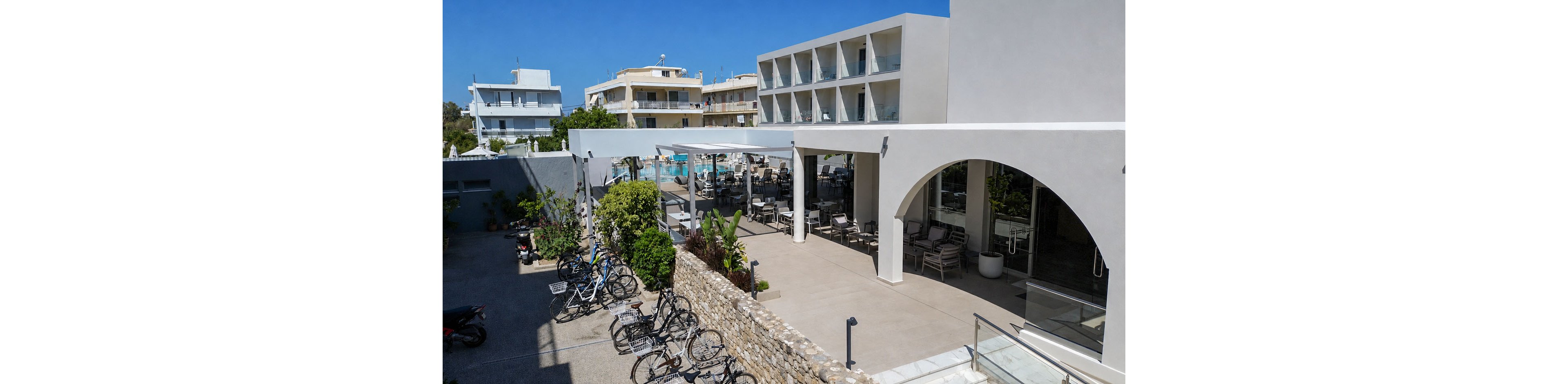 A contemporary hotel complex features a bright courtyard with a shaded terrace and outdoor seating. Several bicycles are parked in the foreground near a stone wall and landscaped plants. In the background, a swimming pool area and neighboring buildings are visible under a clear blue sky. The architecture is minimalist with white facades, clean lines, and glass balconies, creating a relaxed vacation atmosphere.