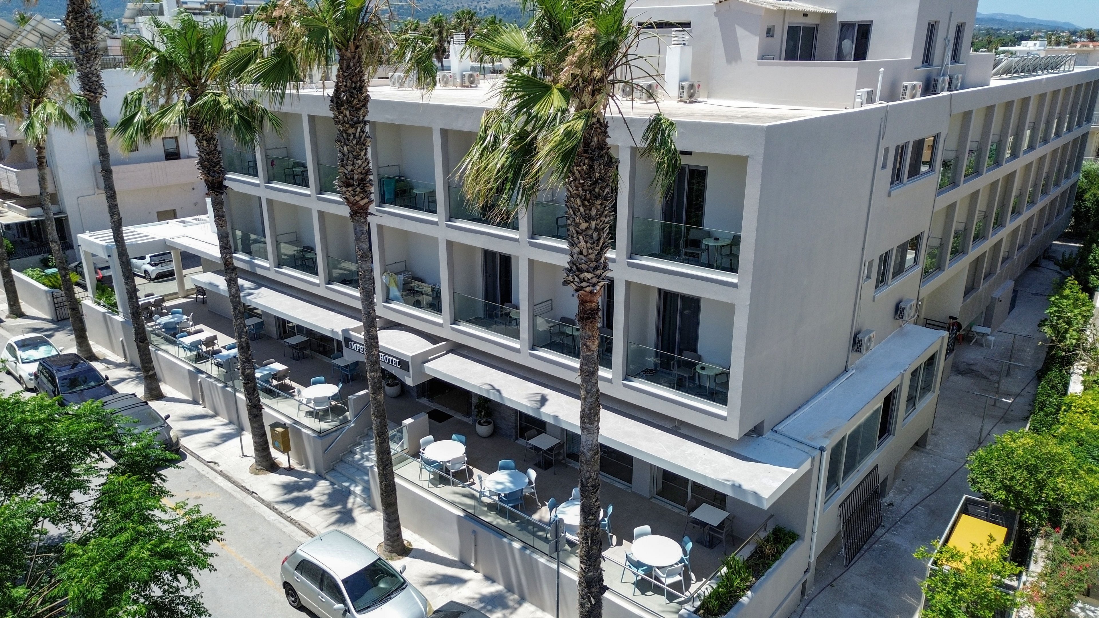 A contemporary multi-story hotel building is shown from an elevated angle on a sunny day. The structure features long rows of balconies and large windows overlooking a street lined with tall palm trees. On the ground level, an outdoor terrace area is set with round tables and chairs, suggesting a café or restaurant space. Parked cars and surrounding greenery indicate an urban coastal setting with relaxed vacation vibes.