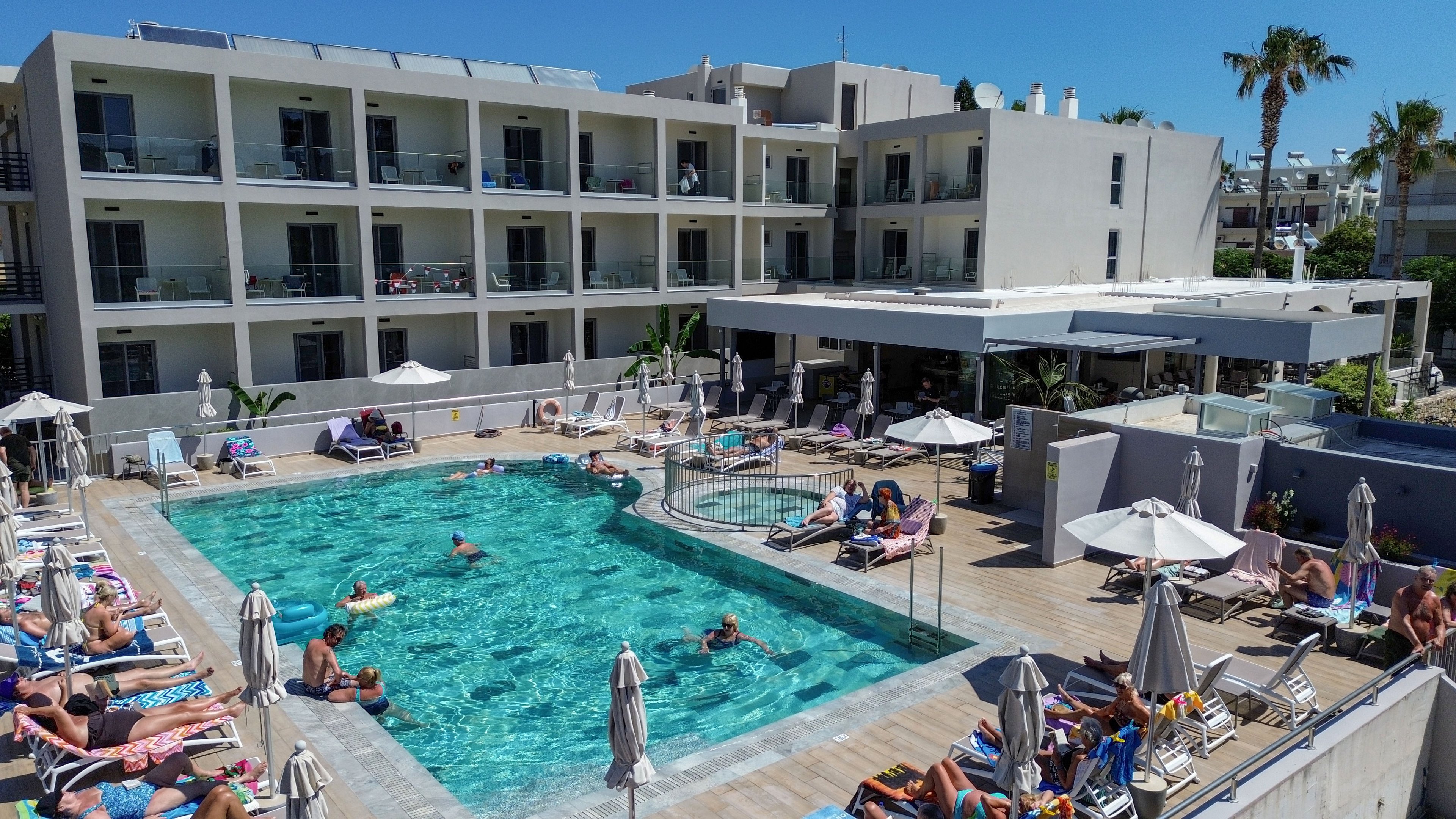 A contemporary multi-story hotel surrounds a large outdoor swimming pool filled with guests enjoying the water. Sunbathers relax on lounge chairs around the pool deck under clear blue skies, with umbrellas providing shade. The building features balconies on each floor, and a smaller circular pool or hot tub is adjacent to the main pool. Palm trees and clean architectural lines create a bright, vacation resort atmosphere.