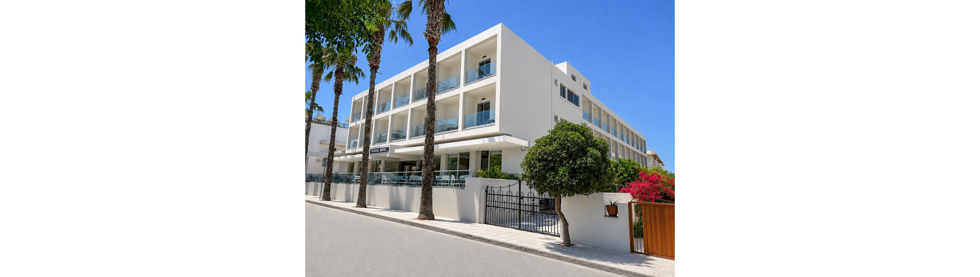 A contemporary white hotel building stands along a quiet street lined with tall palm trees. The structure features multiple floors with balconies and glass railings, giving it a clean, minimalist look. A black metal gate and low wall define the entrance area, with greenery and flowering shrubs adding color to the scene. Bright blue sky and strong sunlight suggest a warm, coastal or Mediterranean climate.