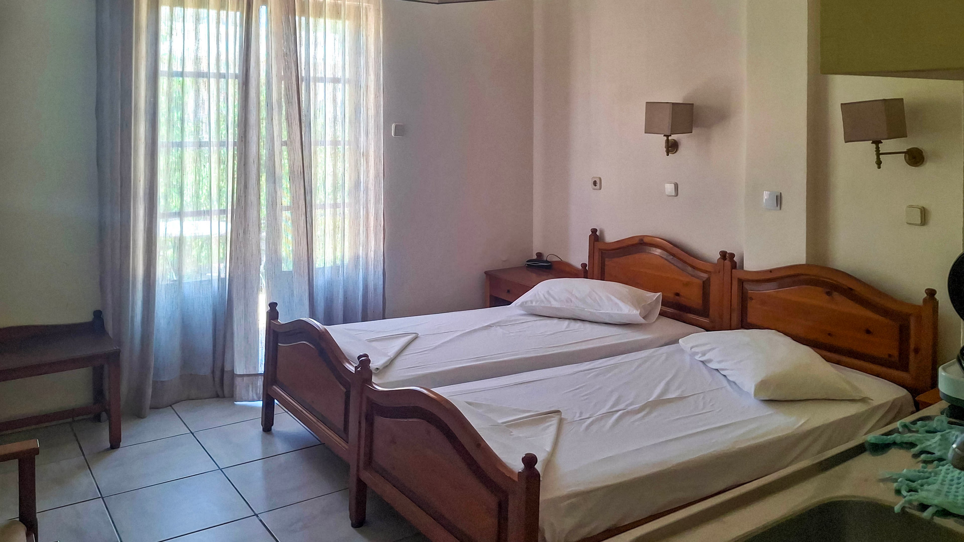 A simple hotel room features two wooden twin beds pushed together with white linens and pillows. Natural light filters through sheer curtains covering a tall window or balcony door, brightening the tiled floor. Wall-mounted bedside lamps and a small nightstand sit between the beds, while a wooden bench stands near the window. The overall atmosphere is clean, minimal, and inviting, suggesting a budget-friendly vacation stay.