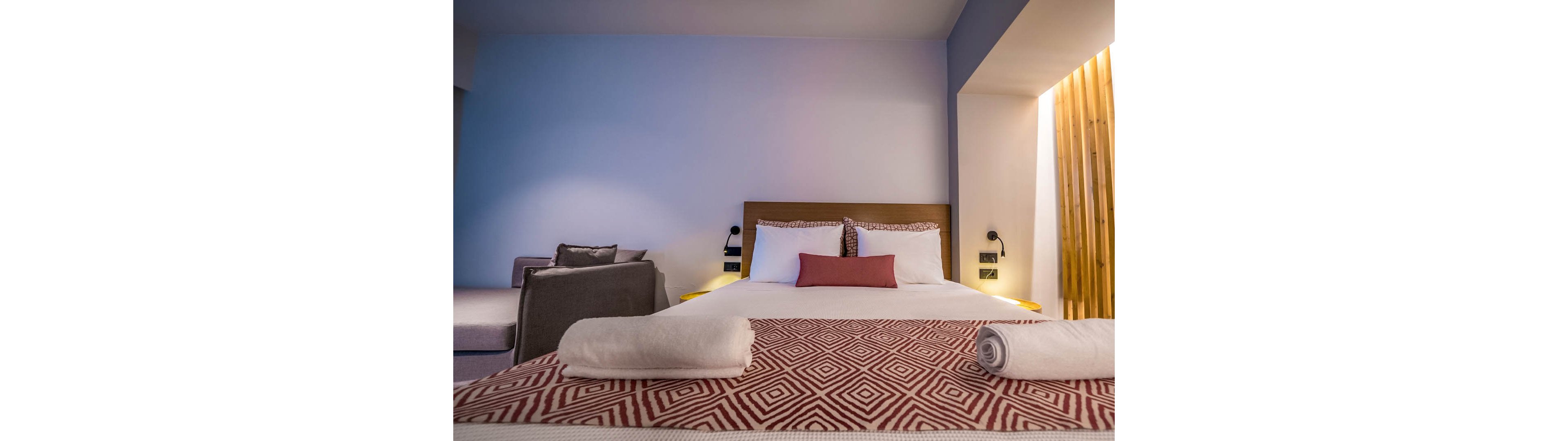 A neatly made double bed with geometric patterned bedding is the main focus in this modern hotel room. The setting features soft lighting, a cozy armchair, and a minimalist design with wooden accents. Two rolled towels are placed at the foot of the bed, and the color palette includes muted tones with pops of red and beige. The overall mood is inviting and tranquil, ideal for relaxation.
