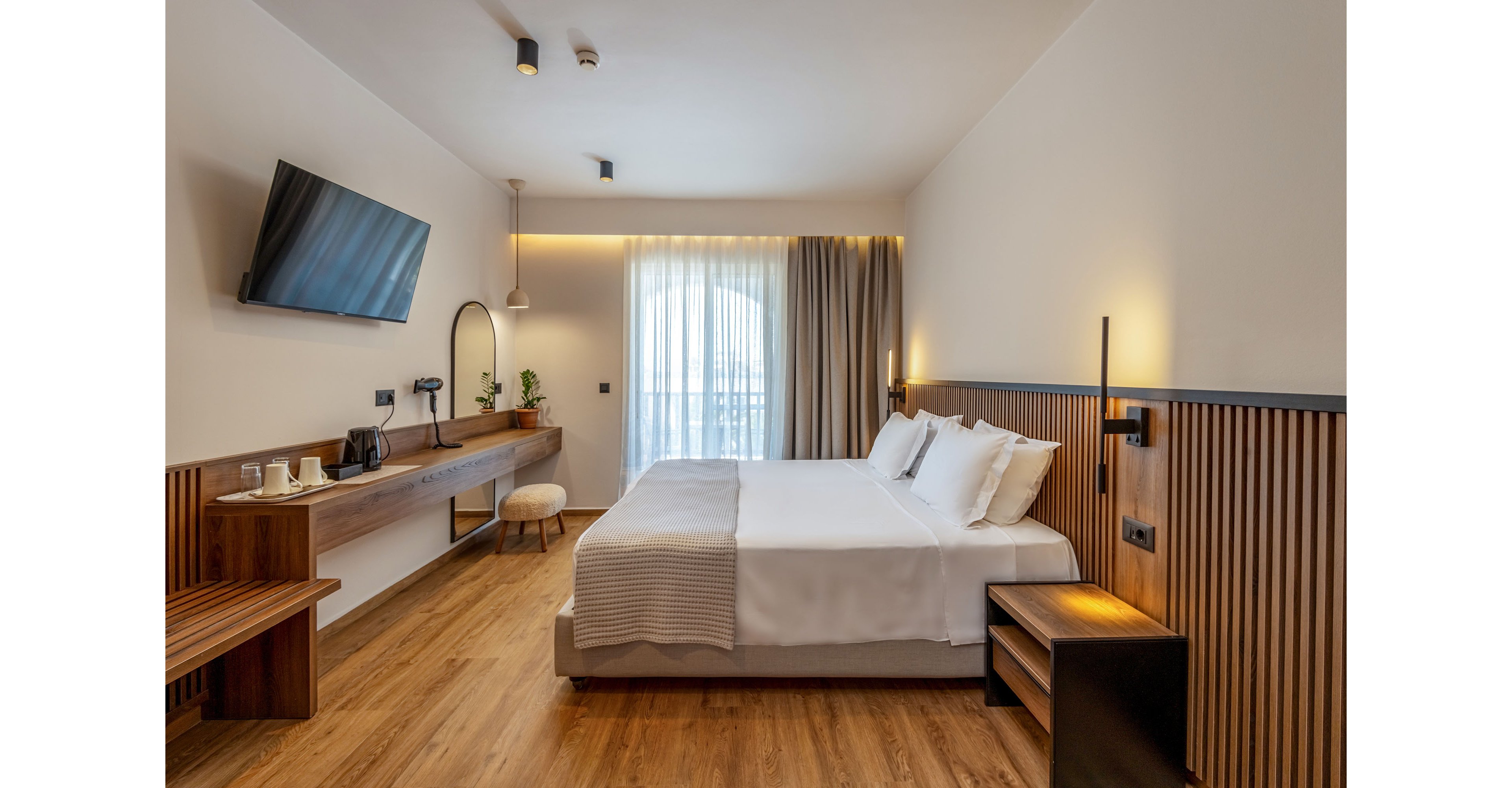 Contemporary hotel room featuring a large double bed with white linens and multiple pillows. The space includes a wall-mounted flat-screen TV, a long wooden desk with a mirror, and a small stool. Floor-to-ceiling window with sheer curtains and blackout drapes lets in natural light, while warm accent lighting creates a cozy atmosphere. Wooden flooring and slatted wood wall panels add a minimalist, modern design touch.