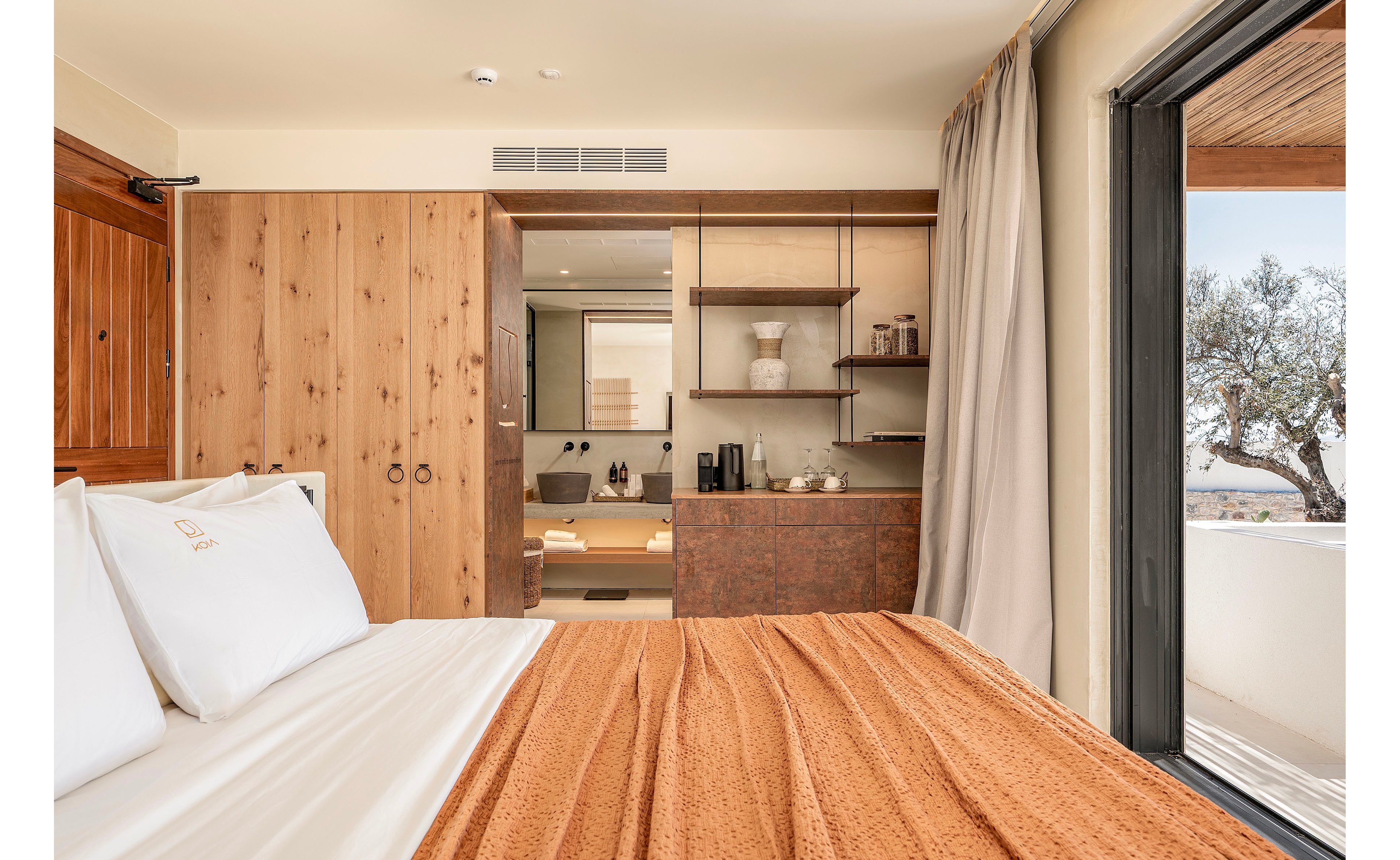 A contemporary hotel bedroom features a neatly made bed with white linens and a textured orange blanket. Natural wood wardrobes and cabinetry frame the view toward an open ensuite bathroom with a freestanding bathtub and double sinks. Shelving with decorative objects and a small beverage station add functional details to the space. Large windows and neutral tones create a warm, inviting atmosphere with abundant natural light.