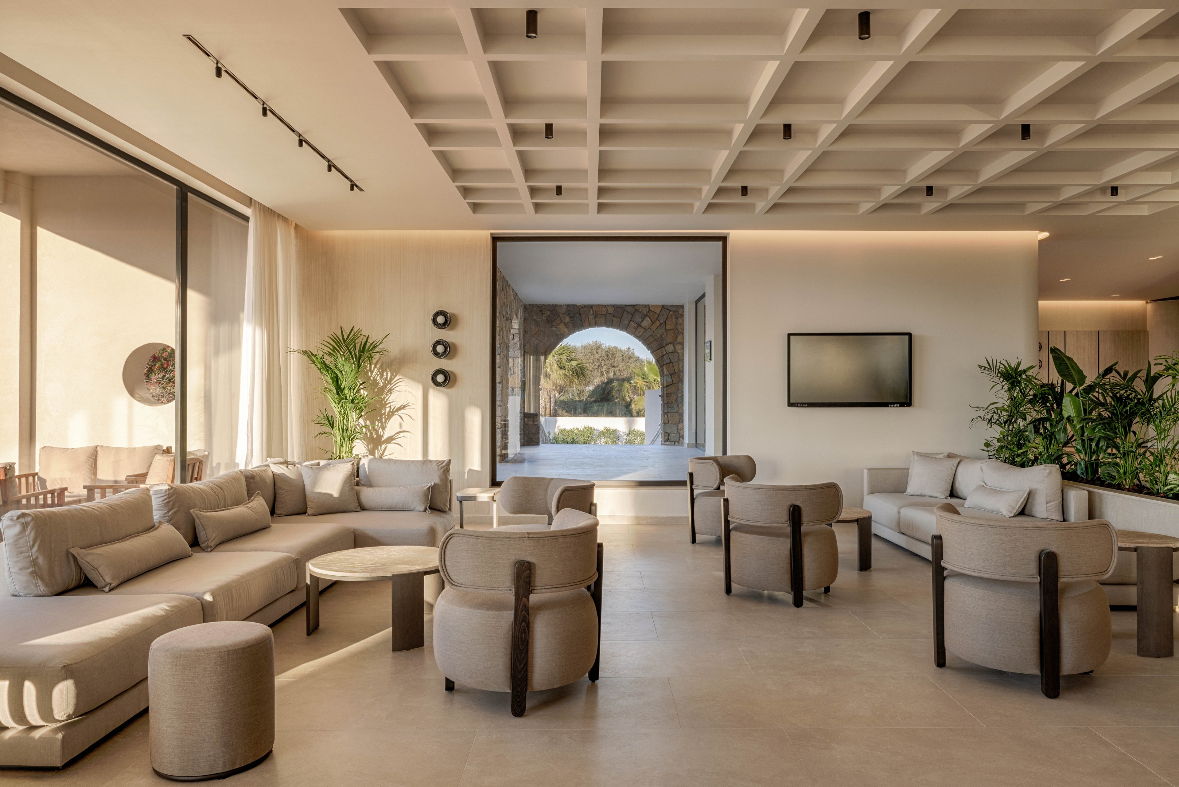 A spacious contemporary lounge features beige sofas and rounded armchairs arranged for relaxed conversation. Large windows and sliding glass doors let in abundant natural light and reveal an outdoor seating area. A wall-mounted television, potted plants, and minimalist decor contribute to the clean, modern aesthetic. The neutral color palette and soft furnishings create a calm, inviting atmosphere suitable for hospitality or residential settings.