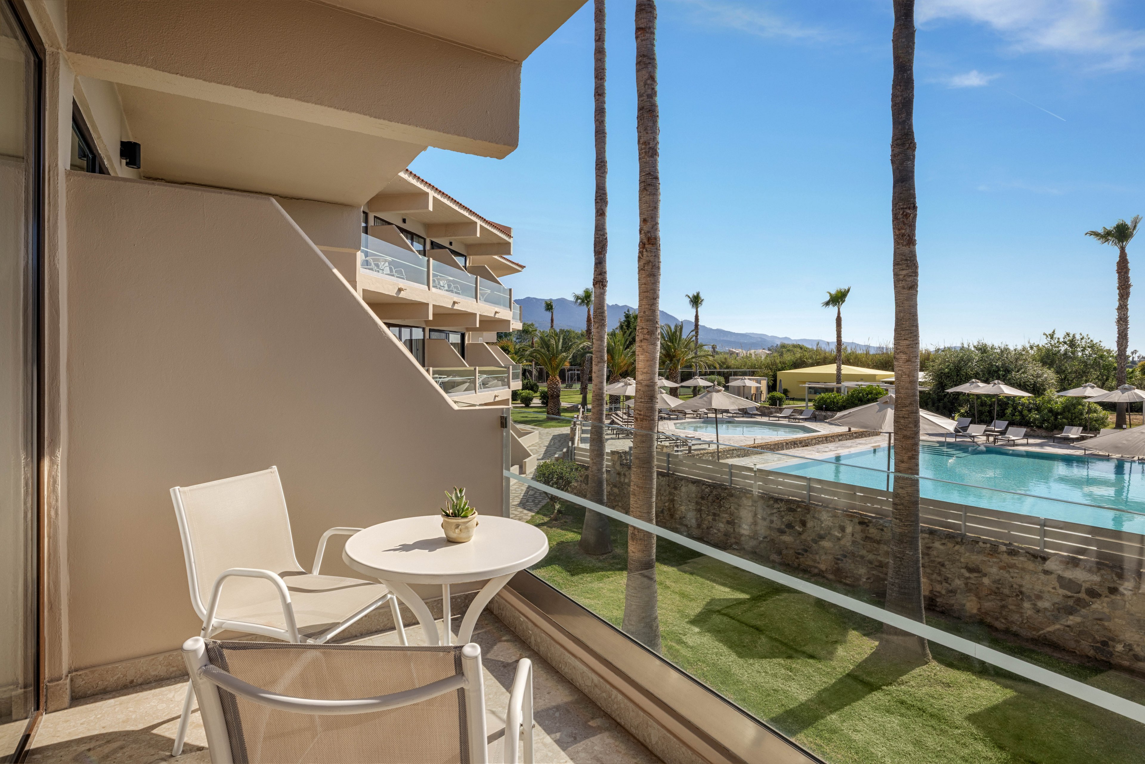 A small hotel balcony features a round white table and two chairs, with a potted plant adding a touch of greenery. The balcony looks out over a large outdoor swimming pool surrounded by sun loungers and umbrellas. Tall palm trees line the pool area, and modern resort buildings frame the scene. In the distance, low mountains and a clear blue sky suggest a warm, sunny vacation destination.