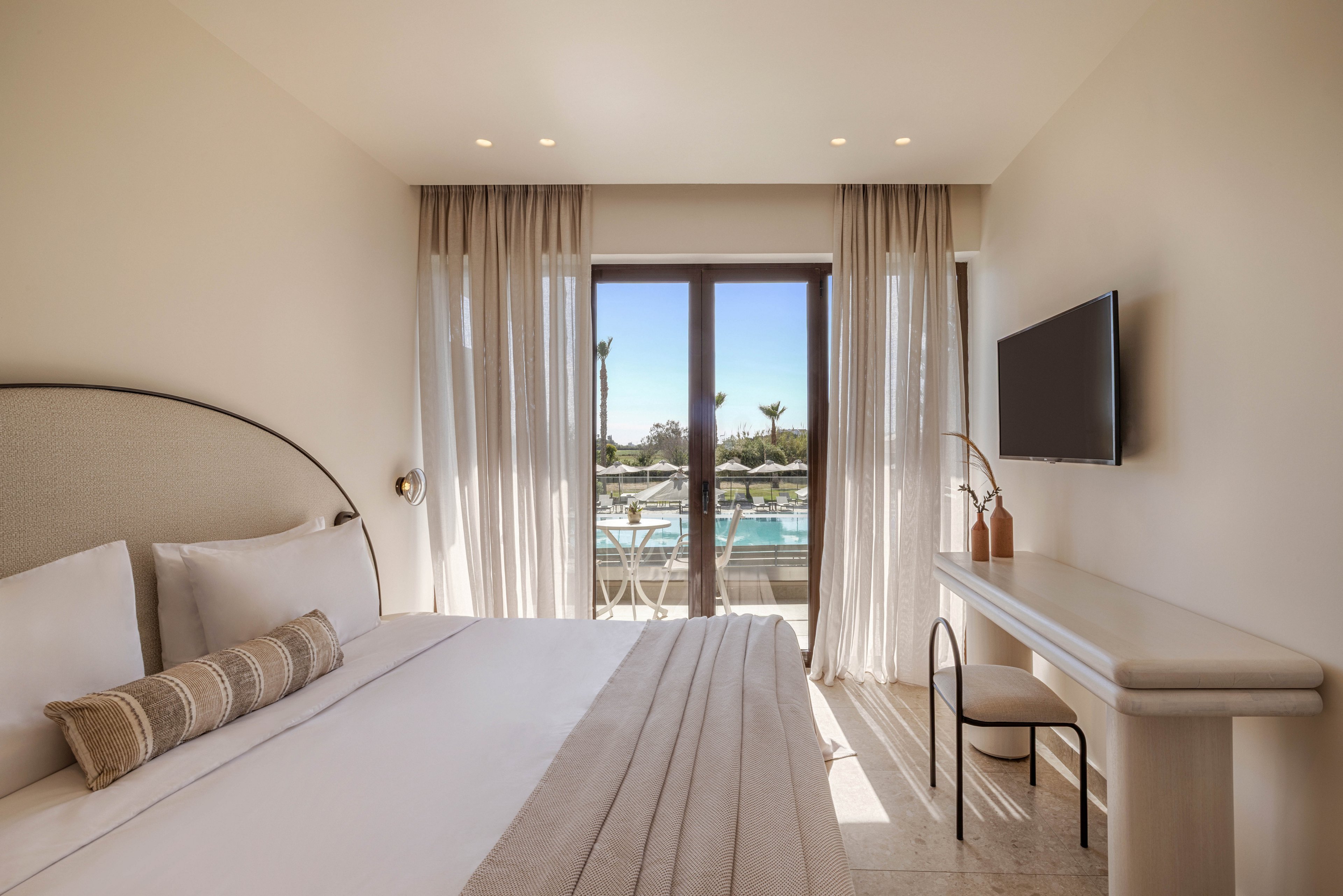 A contemporary hotel bedroom features a neatly made bed with neutral linens and a decorative pillow. Sheer curtains frame large glass doors that open to a balcony overlooking a swimming pool and palm trees. A wall-mounted television faces the bed, and a minimalist desk with two stools sits along the opposite wall. Soft, warm lighting and beige tones create a calm, relaxing atmosphere.