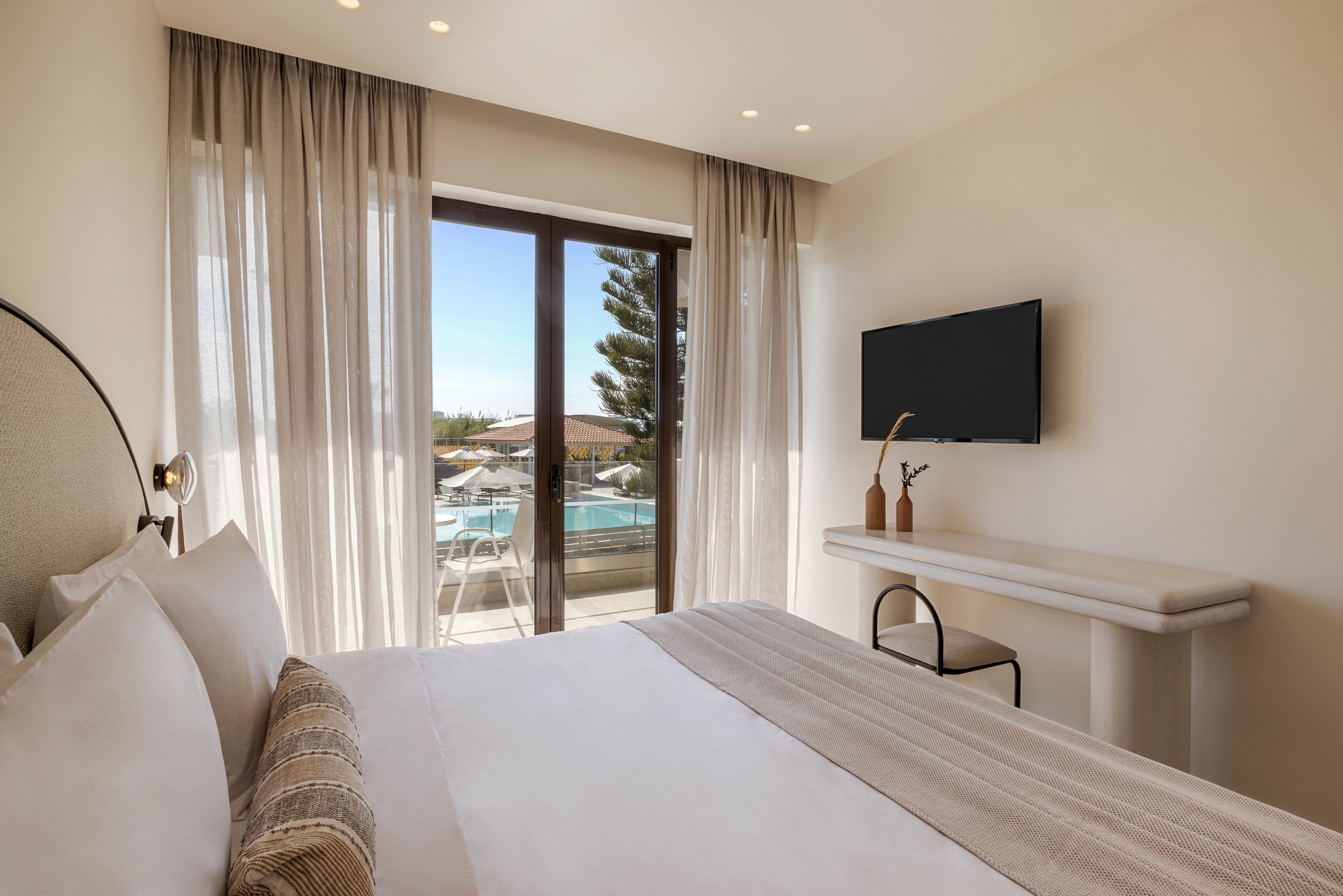 A modern hotel bedroom features a neatly made bed with neutral-toned linens and pillows. Floor-to-ceiling sheer curtains frame a glass door that opens to a balcony overlooking a swimming pool and surrounding buildings. A wall-mounted flat-screen TV faces the bed above a minimalist white desk with decorative vases and a chair. Soft recessed ceiling lights and a calm beige color palette create a serene, contemporary atmosphere.