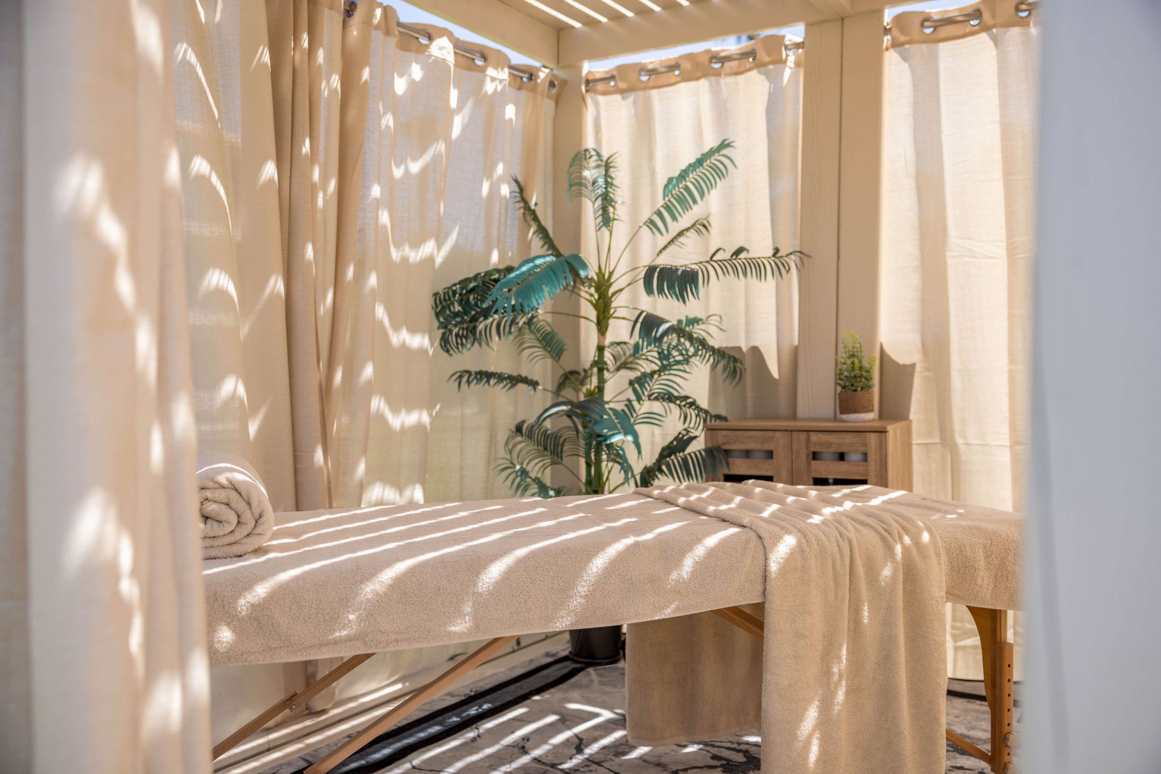A massage table covered in soft beige linens sits inside a sunlit outdoor cabana. Light filters through slatted panels and curtains, casting striped shadows across the table and floor. A rolled towel rests at one end of the table, ready for a spa treatment. Potted plants and a small wooden cabinet add a calming, natural touch to the serene setting.