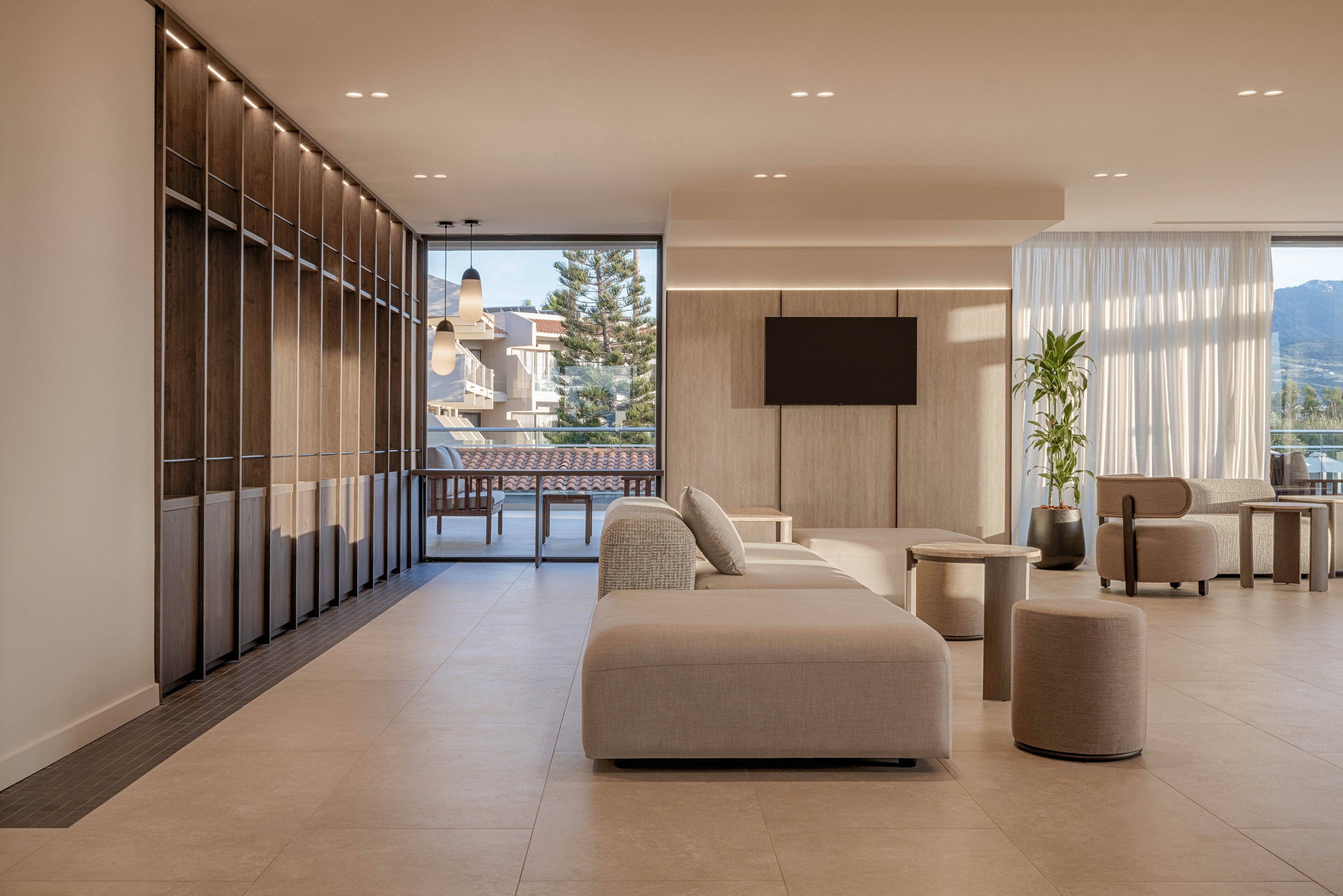 Spacious contemporary living room featuring low-profile beige sofas and ottomans. Large floor-to-ceiling windows open to a balcony with views of trees and distant mountains. Warm wood paneling, built-in shelving, and a wall-mounted television create a clean, minimalist aesthetic. Soft neutral tones and indirect lighting contribute to a calm, luxurious atmosphere.