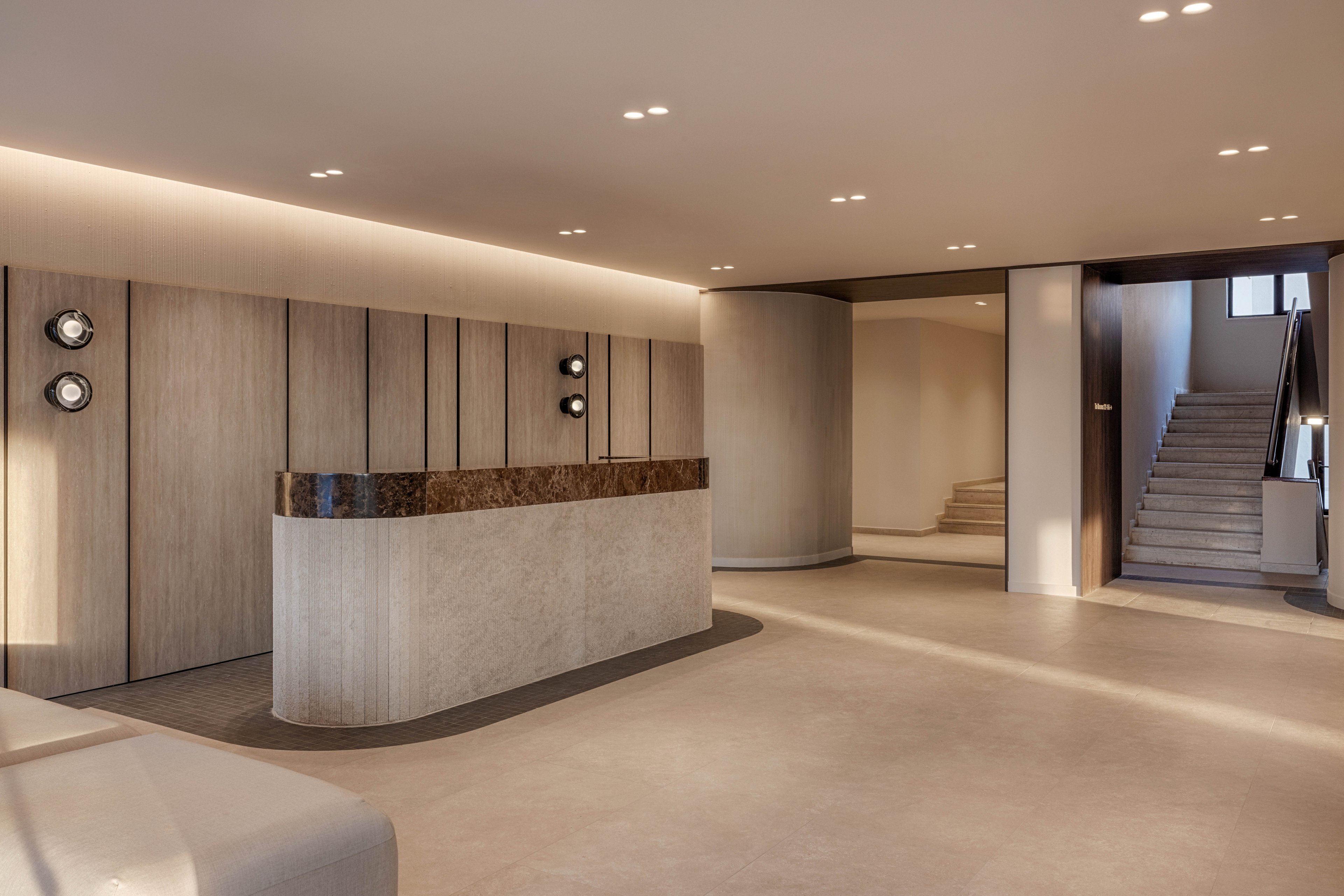 A contemporary lobby features a sleek reception desk with a stone base and dark countertop. Vertical wood panels line the wall behind the desk, accented by circular wall lights. Recessed ceiling lights softly illuminate the spacious, uncluttered interior. In the background, a staircase and hallway lead to other areas of the building, enhancing the sense of depth and openness.
