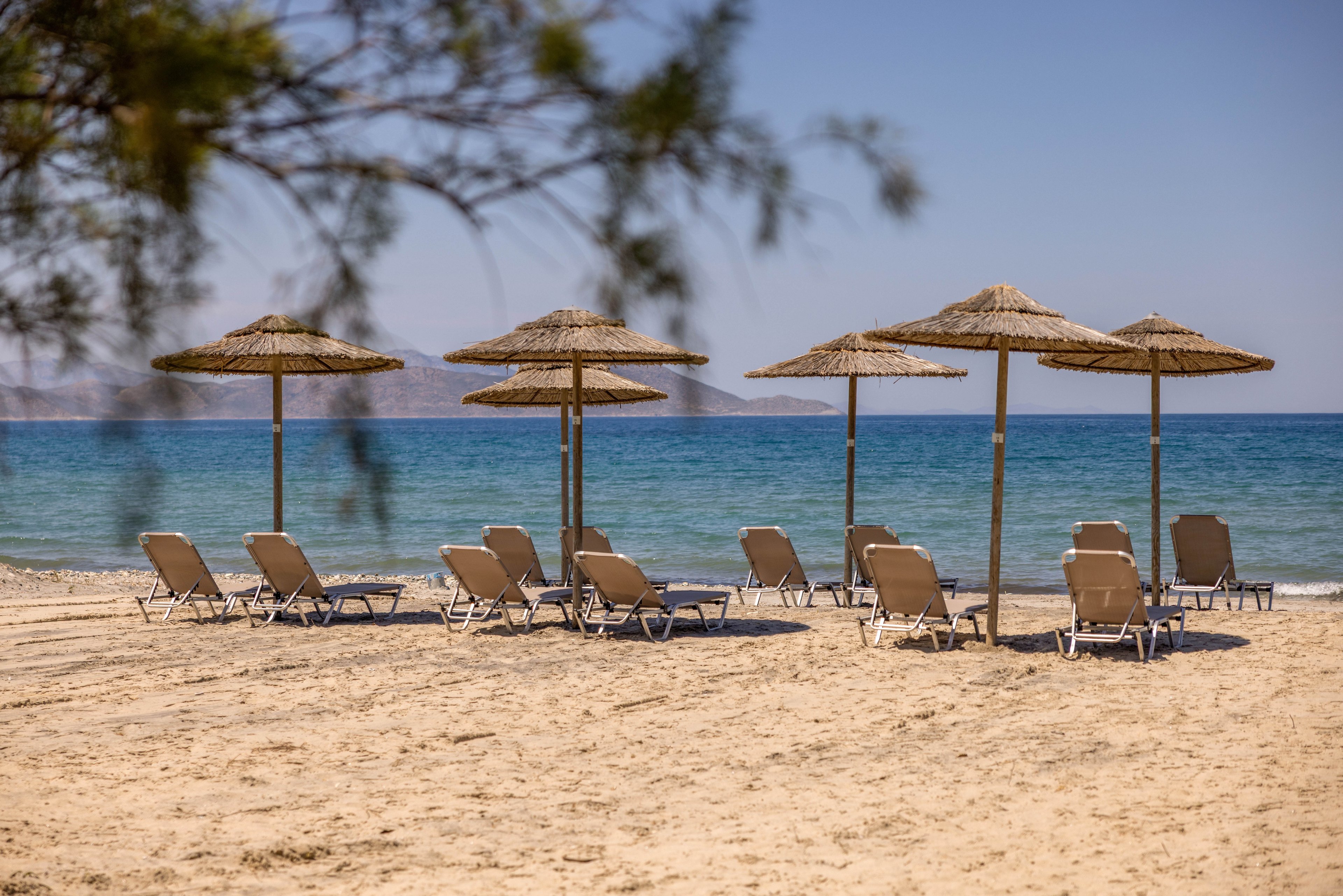 A row of empty sun loungers sits beneath straw parasols on a quiet sandy beach. Calm blue sea stretches out to the horizon under a clear sky. Distant landforms are faintly visible across the water, suggesting a peaceful coastal destination. The scene evokes relaxation and a laid-back summer holiday atmosphere.
