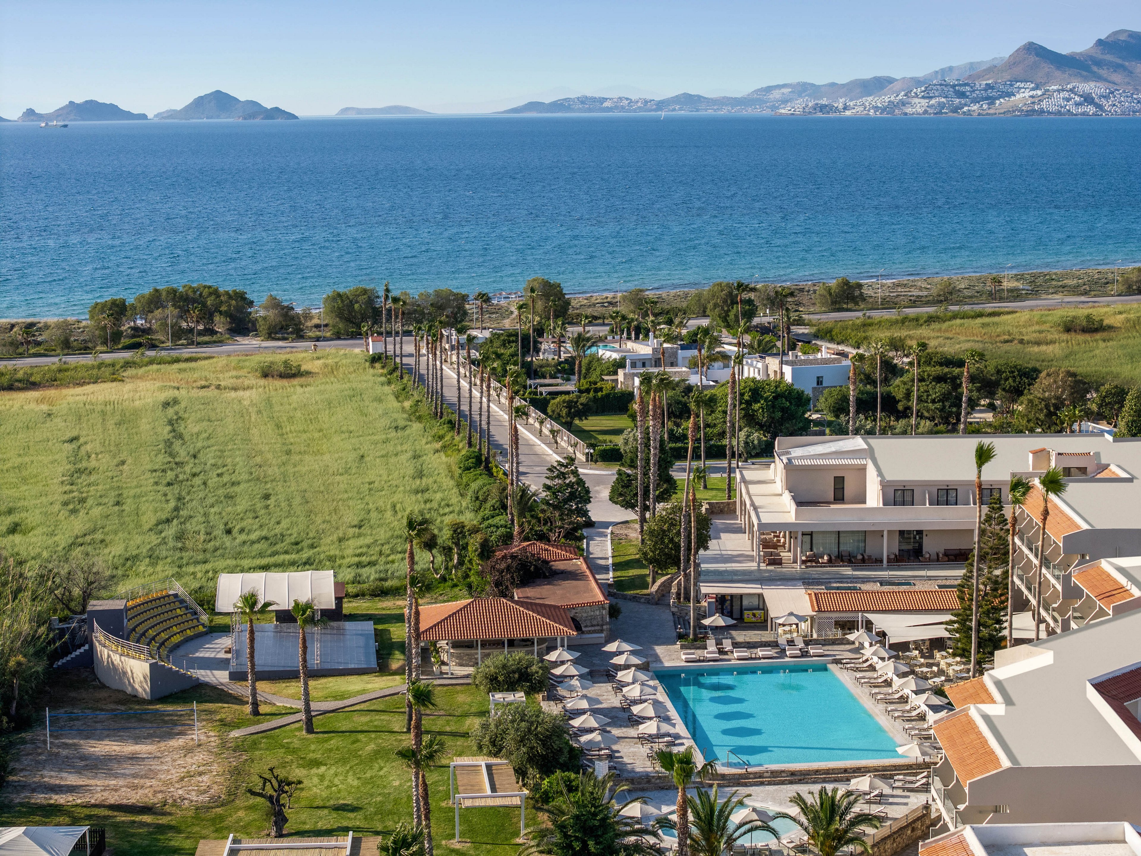 A seaside resort complex features a central outdoor swimming pool surrounded by sun loungers and palm trees. The property sits beside a wide grassy field that stretches toward a coastal road and the shoreline. Calm blue water extends to distant mountains and a town across the bay under a clear sky. The scene suggests a peaceful vacation destination with ample outdoor space and scenic ocean views.