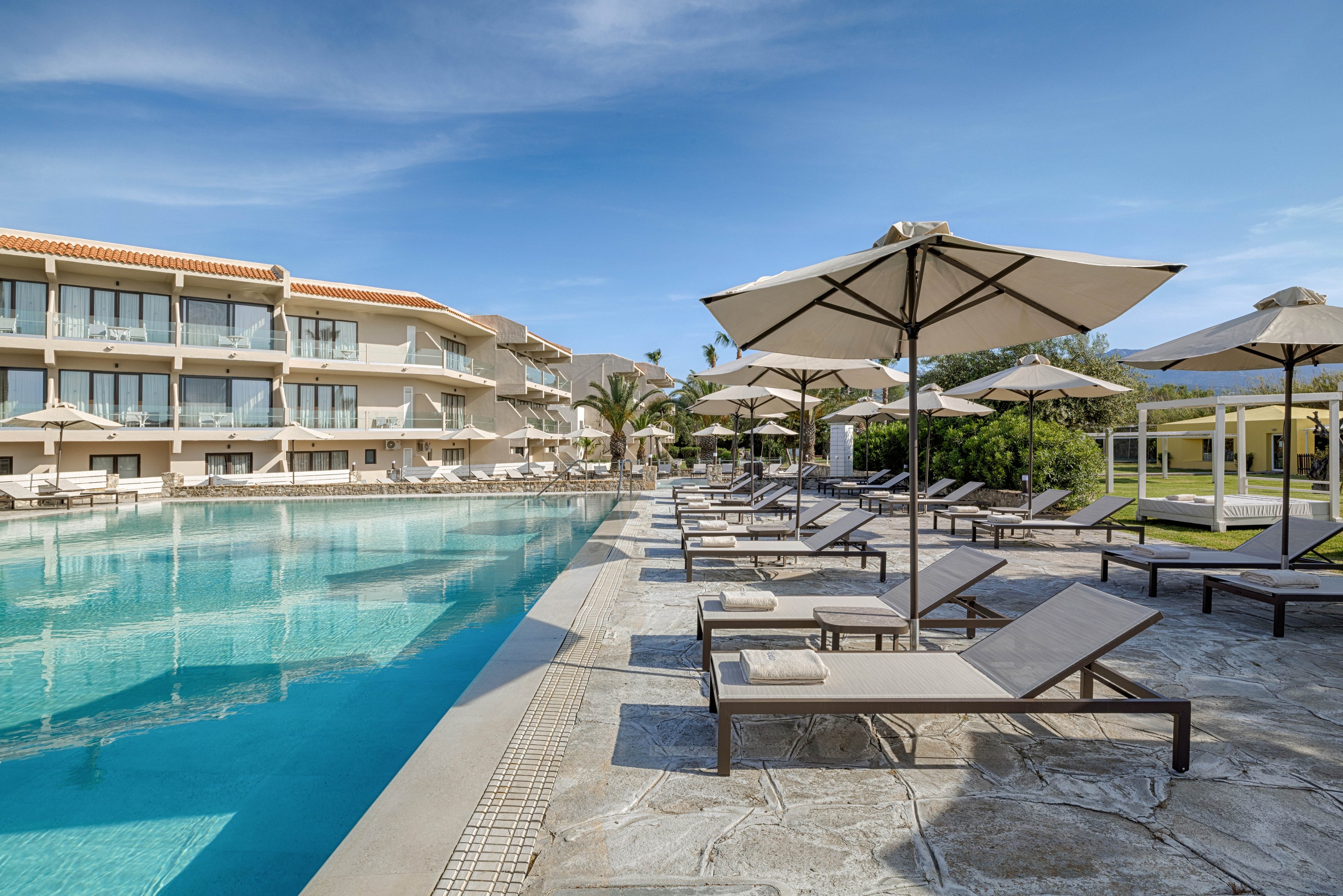 A large outdoor swimming pool stretches alongside a modern resort building with balconies. Numerous sun loungers and umbrellas line the stone pool deck, all currently unoccupied. Palm trees and manicured greenery add a tropical touch to the scene. The clear blue sky and calm water create a relaxed vacation atmosphere.
