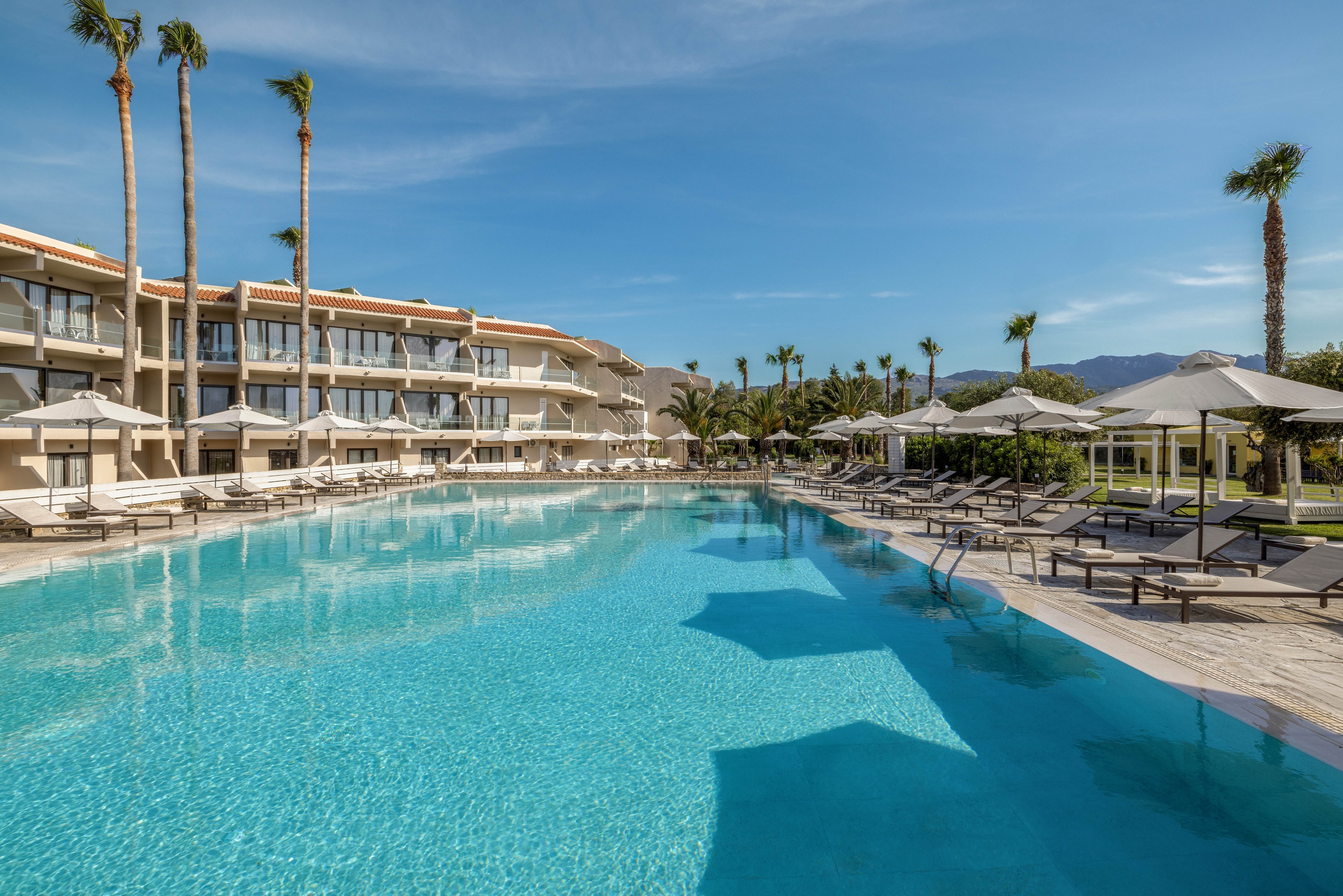 A large, clear swimming pool stretches alongside a modern resort building with balconies. Numerous sun loungers and umbrellas line the wooden deck, suggesting a relaxing vacation setting. Tall palm trees surround the area under a bright blue sky with light clouds. Distant mountains are visible in the background, enhancing the scenic, tranquil atmosphere.