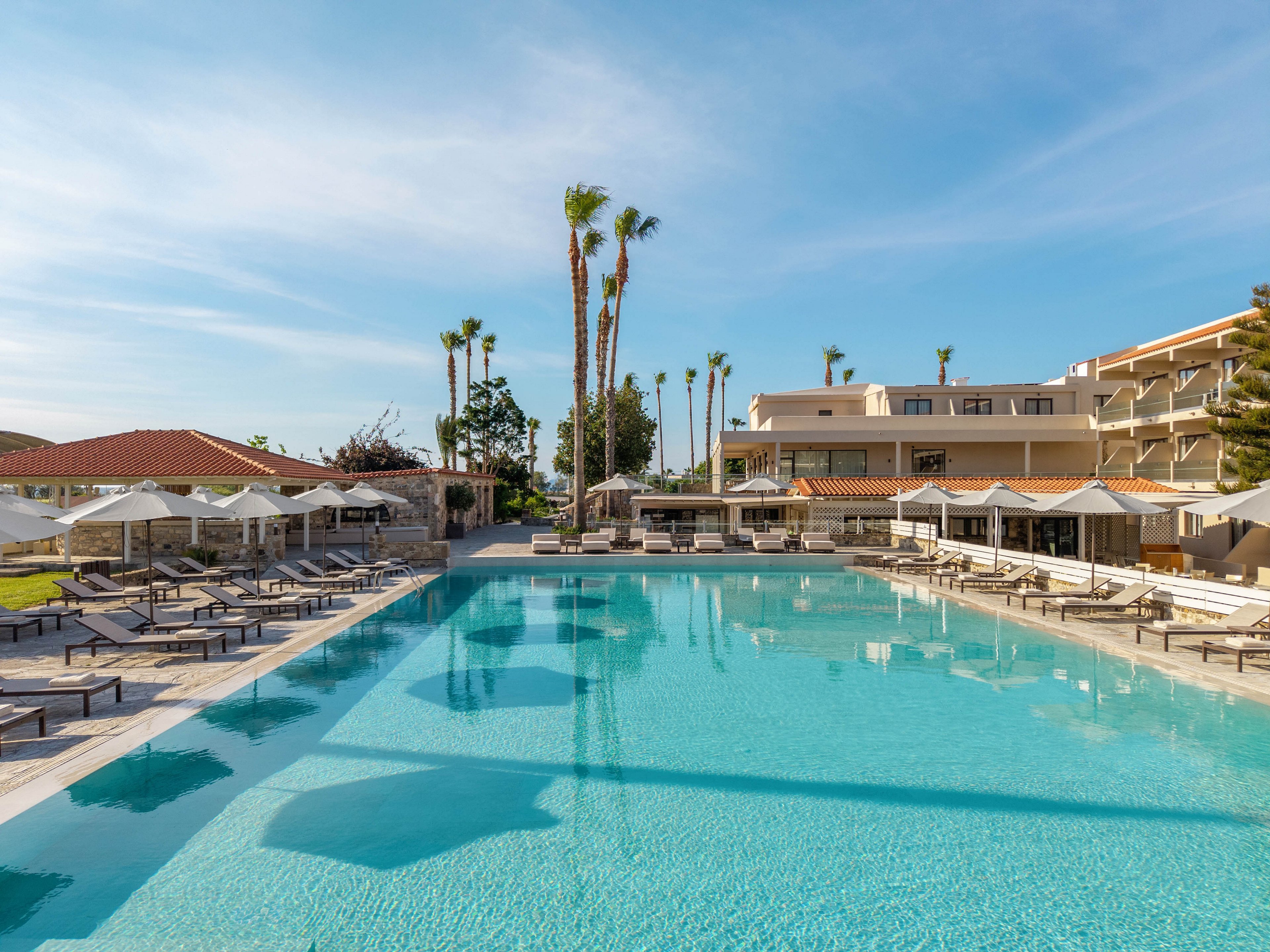 A large, clear blue swimming pool stretches across the foreground of a modern resort. Numerous sun loungers and white umbrellas line both sides of the pool, suggesting a relaxing vacation setting. Multi-story hotel buildings and palm trees rise in the background under a bright blue sky. The scene conveys a calm, sunny atmosphere ideal for leisure and holidays.