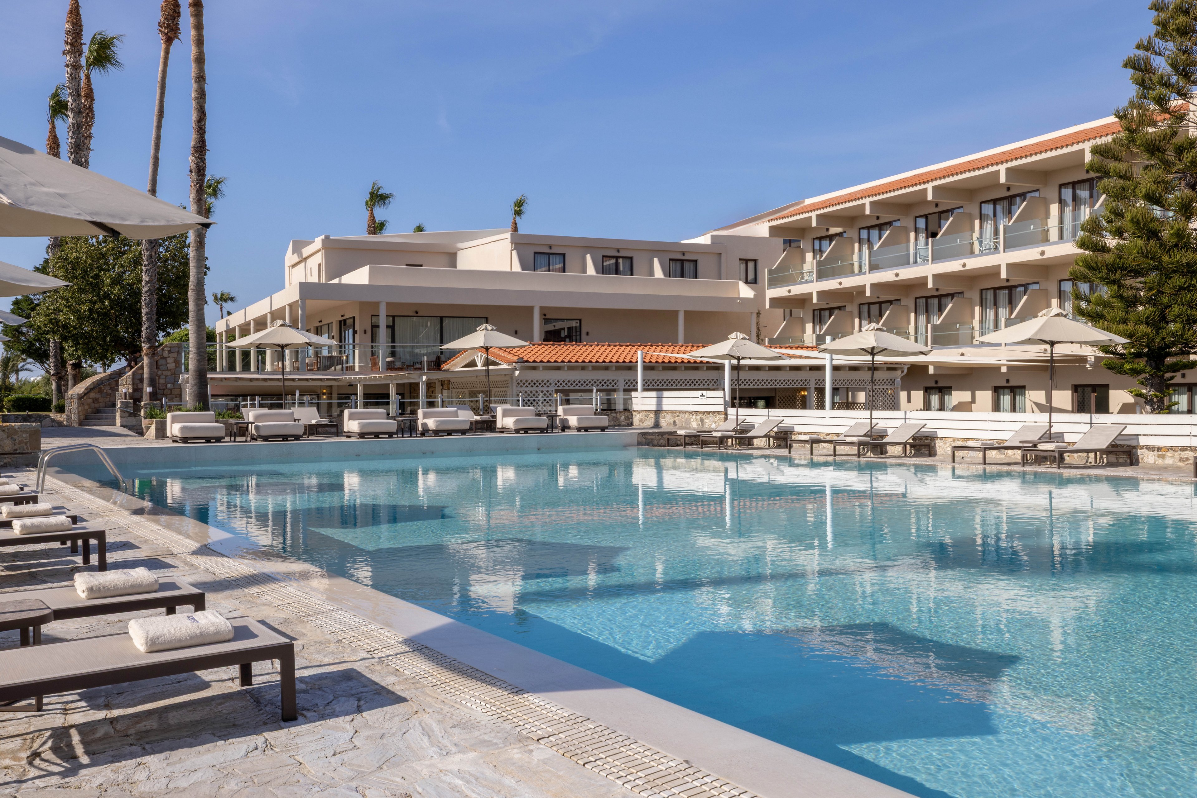 A modern resort hotel features a large, clear blue outdoor swimming pool in the foreground. Sun loungers and umbrellas line the poolside terrace, suggesting a relaxing vacation setting. The multi-story hotel buildings with balconies overlook the water, framed by tall palm trees and a bright blue sky. The scene conveys a calm, sunny, and luxurious holiday atmosphere.