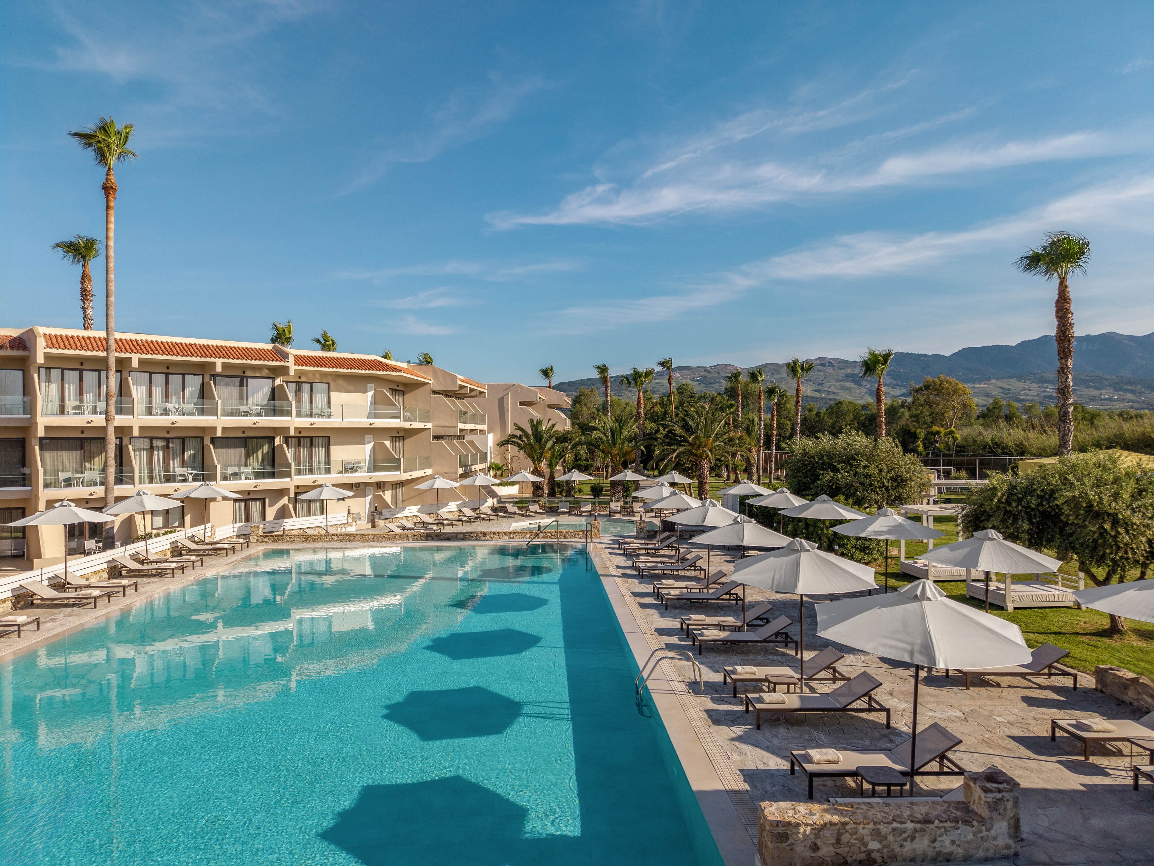 A contemporary resort hotel features a large, clear blue outdoor swimming pool surrounded by sun loungers and white umbrellas. The multi-story building with balconies overlooks the pool area, offering views of the landscaped grounds. Palm trees and green shrubs line the pathways, adding a tropical feel to the setting. In the distance, low mountains rise under a bright blue sky with light clouds, creating a calm vacation atmosphere.