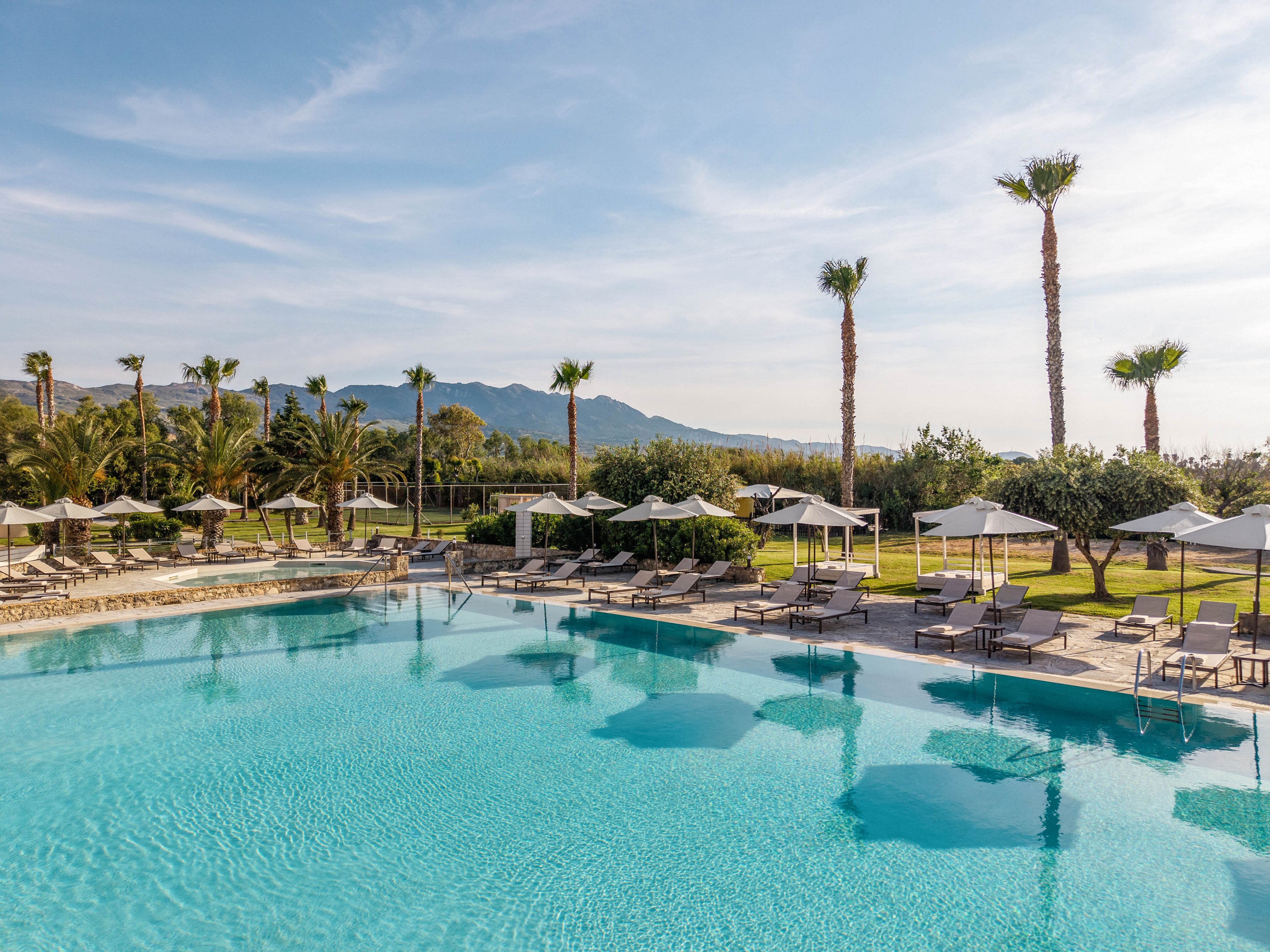 A large, clear blue swimming pool is surrounded by sun loungers and white parasols at an upscale resort. Tall palm trees line the pool area, creating a tropical atmosphere. In the background, green fields and distant mountains are visible under a bright, partly cloudy sky. The scene suggests a calm, sunny day ideal for relaxation and vacation leisure.