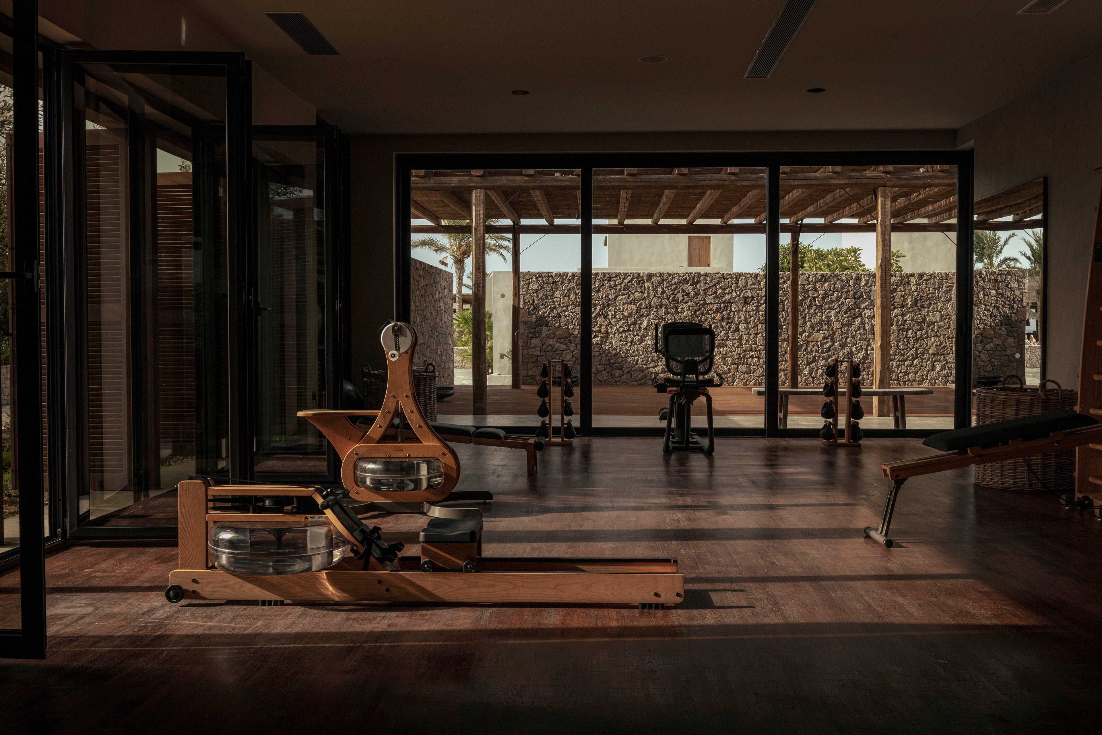 A contemporary fitness room features wooden rowing machines and a stationary exercise bike as its main equipment. The space is illuminated by natural light streaming through large glass doors, creating a warm and inviting atmosphere. The gym overlooks an outdoor patio with stone walls, adding to the tranquil and stylish setting. No visible text or numeric values are present in the image.