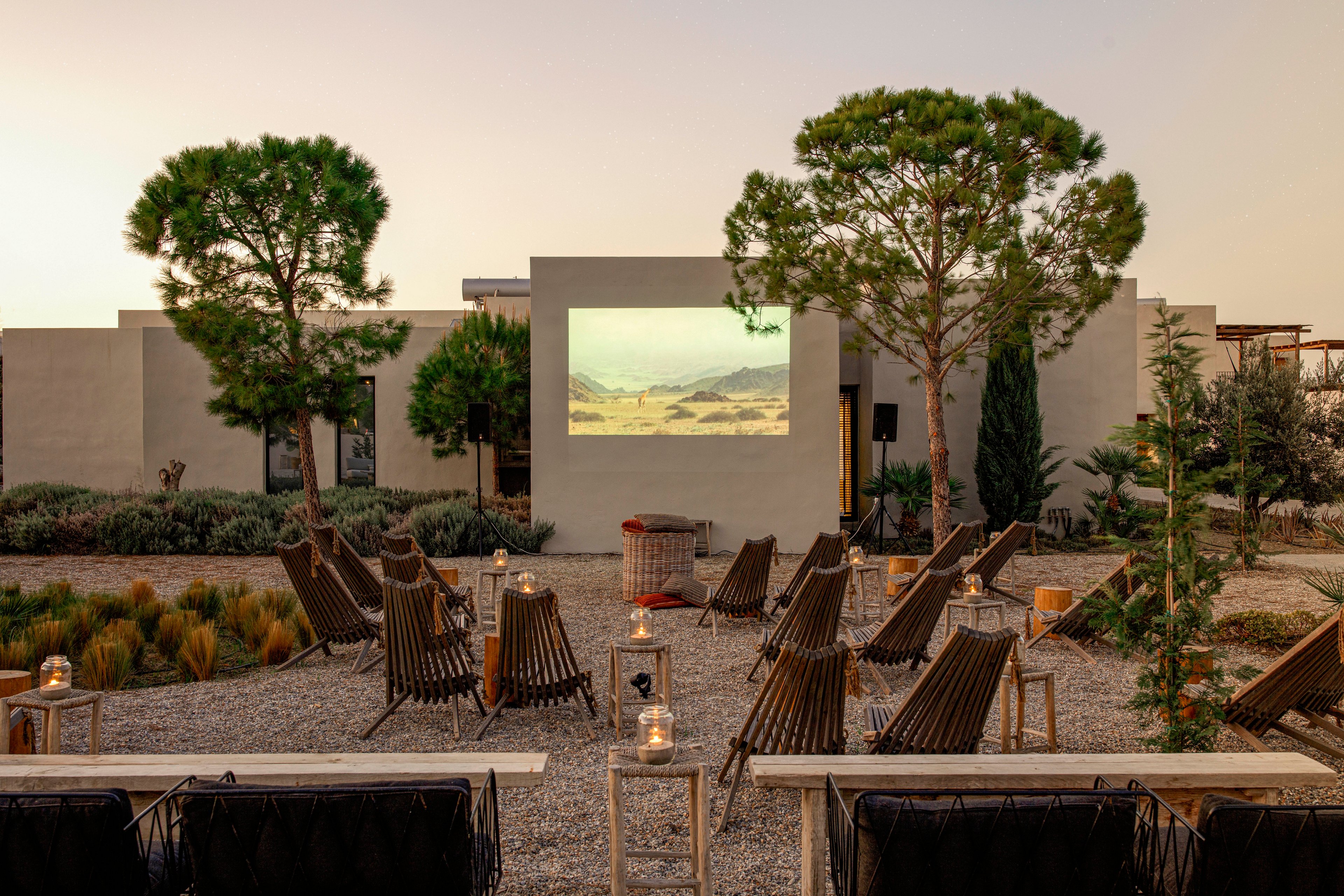 The image shows an outdoor movie screening area with rows of lounge chairs facing a large projection screen. The setting is a landscaped courtyard with gravel ground, trees, and modern architecture in the background. Lanterns and small tables are placed beside the chairs, creating a cozy and inviting atmosphere. The scene is captured during sunset, giving the space a warm and tranquil mood.