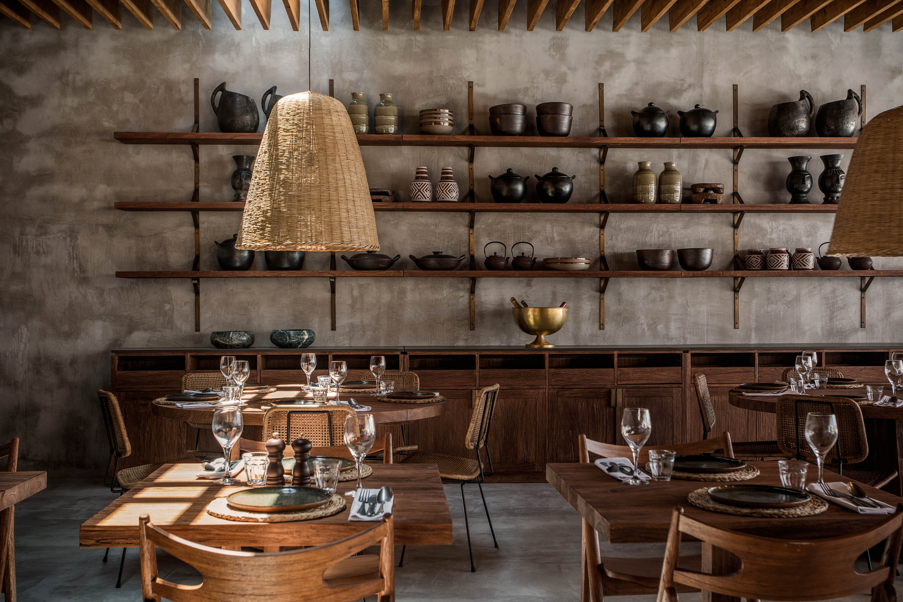The image shows a stylish restaurant dining area featuring wooden tables and chairs, set with glassware and plates. The setting is illuminated by woven pendant lights, with shelves displaying ceramic pots and vases against a textured concrete wall. The overall mood is warm and inviting, with earthy tones and natural materials creating a cozy atmosphere. No visible numeric values or identifiable brands are present.