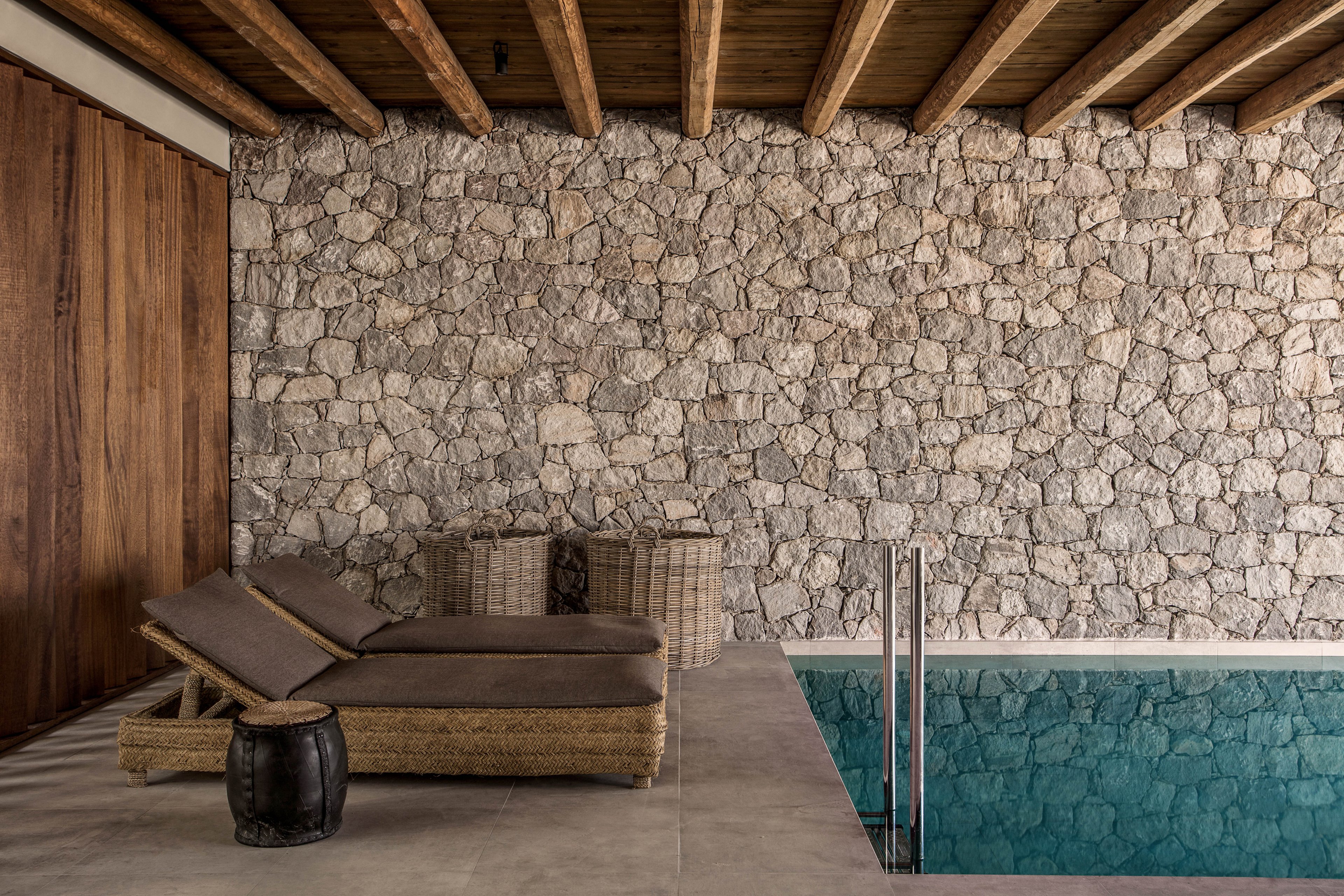 The image features a contemporary indoor pool area with a rustic stone wall and wooden ceiling beams. Two woven lounge chairs with cushions are positioned beside the pool, accompanied by a small dark side table. The setting is tranquil and minimalist, with neutral tones and natural textures creating a relaxing atmosphere. No visible text or numeric values are present in the image.
