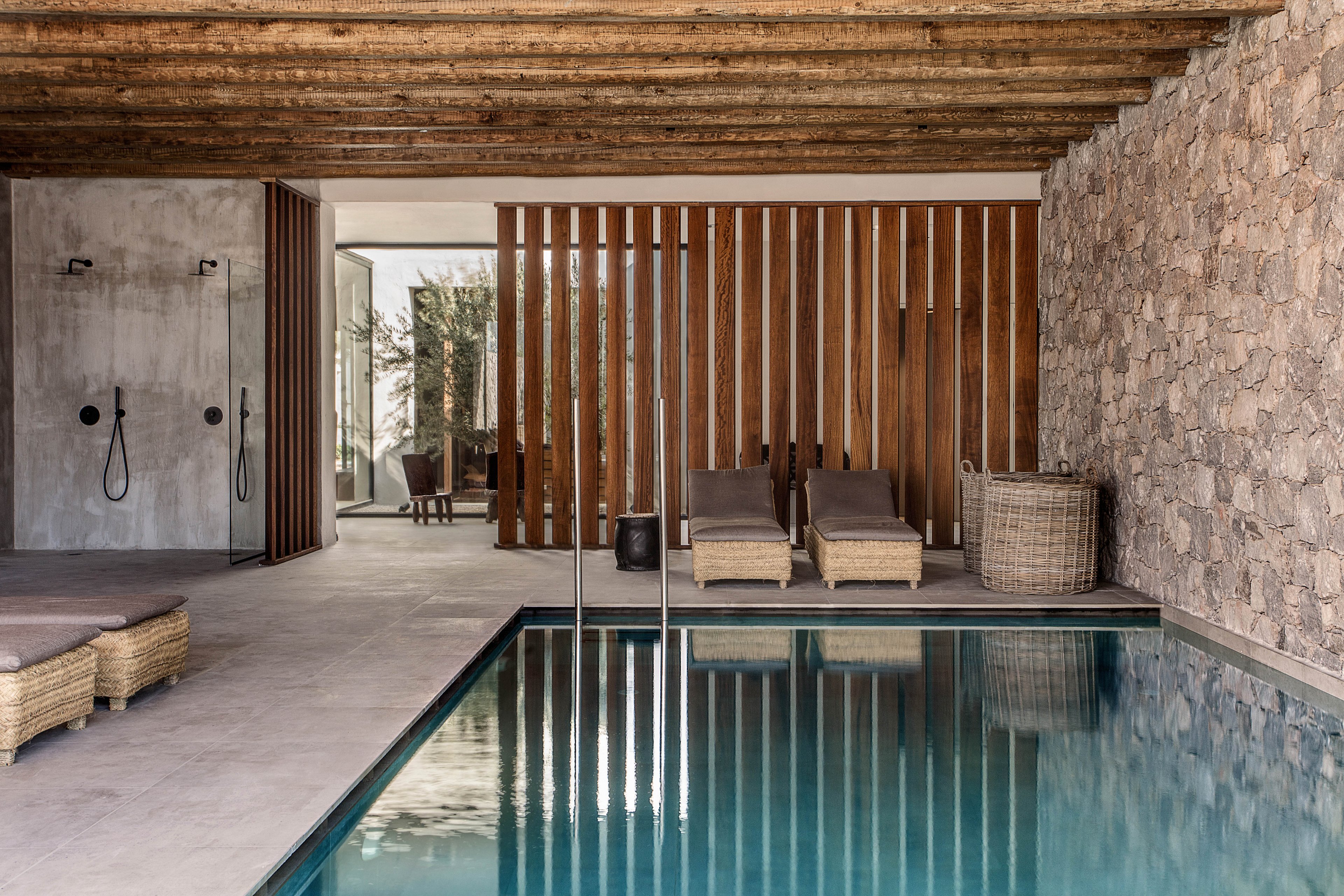The image features a contemporary indoor swimming pool with clear water, surrounded by minimalist decor. Two wicker lounge chairs are positioned near a wooden slat divider, and a large woven basket sits nearby. The setting includes a rustic stone wall, exposed wooden ceiling beams, and an open shower area, creating a serene and natural ambiance. Neutral tones and natural materials dominate the palette, evoking a calm and relaxing mood.