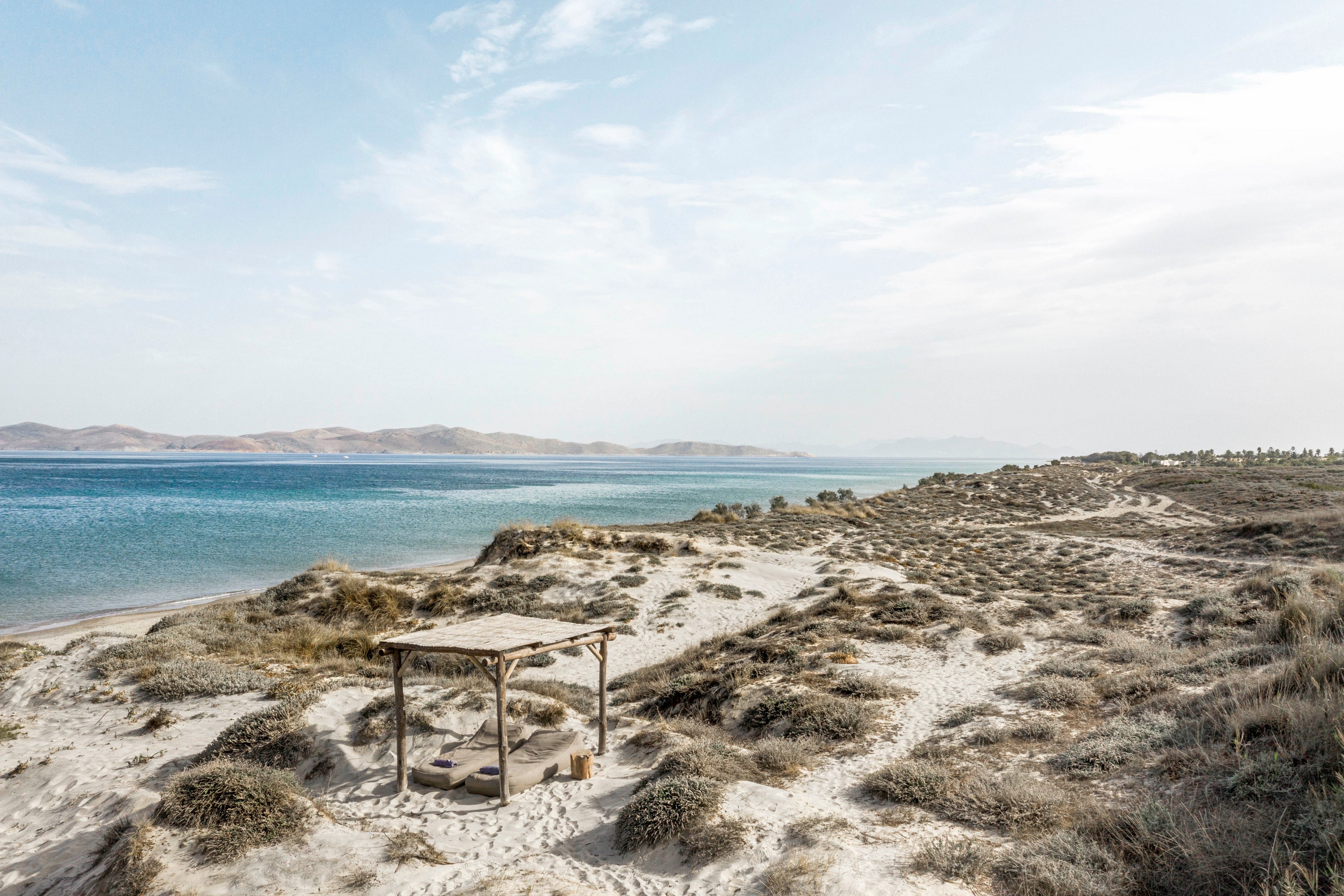 A simple wooden shelter stands on sandy dunes overlooking a tranquil blue sea. The landscape features sparse vegetation and rolling hills in the distance, creating a serene and natural setting. The sky is partly cloudy, and the overall mood is peaceful and remote, with earthy tones dominating the palette. No people or visible text are present in the image.