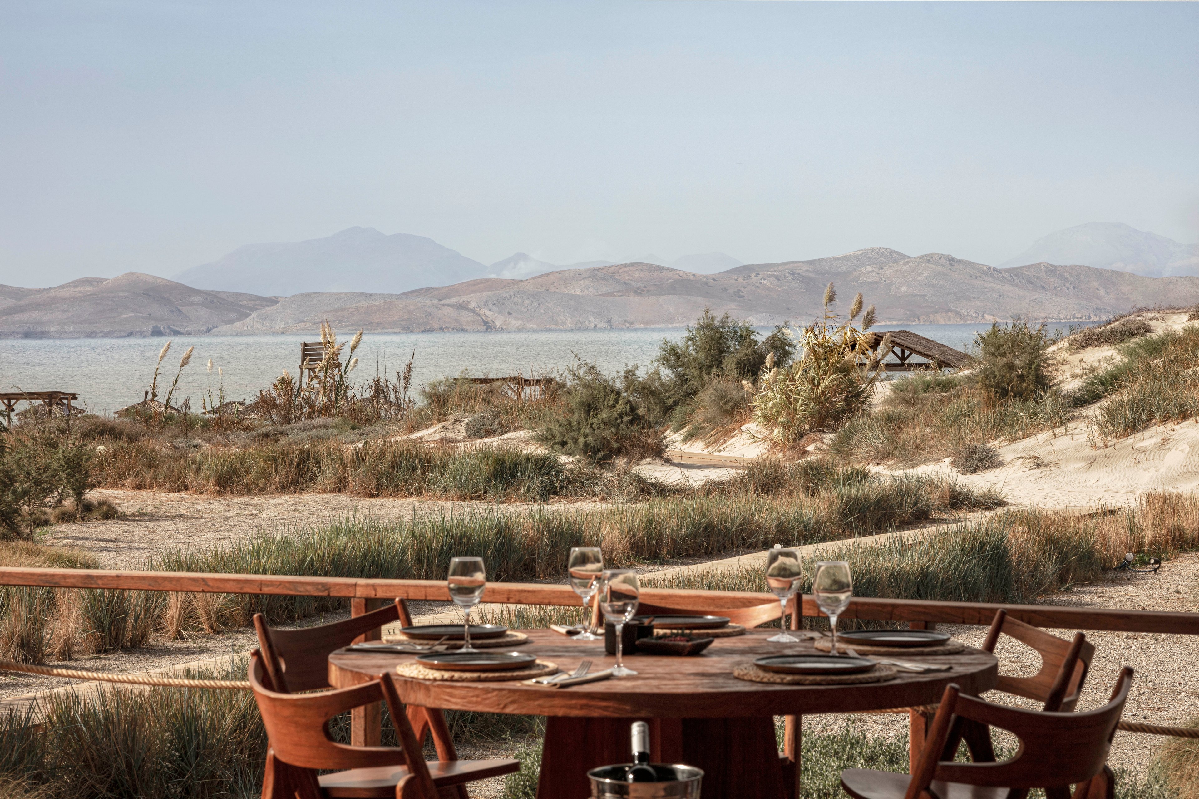 A round wooden dining table is elegantly set with plates, glasses, and cutlery, positioned on a terrace with a scenic view of sandy dunes and calm sea. The setting suggests a relaxed, upscale outdoor meal, with natural light and earthy tones dominating the palette. The background features distant mountains and sparse vegetation, enhancing the tranquil atmosphere. No visible text or numeric values are present in the image.
