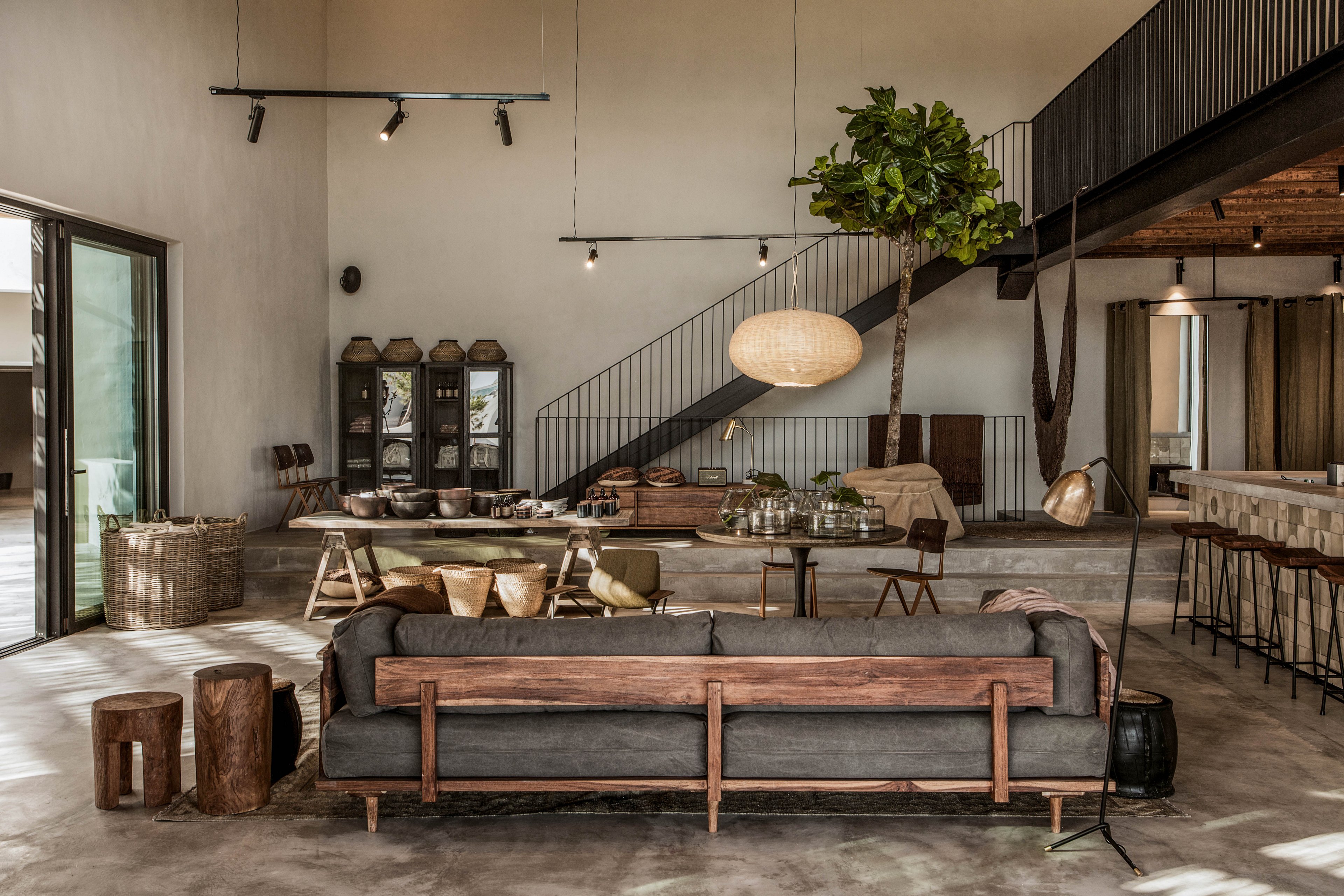 A spacious, contemporary living room features a minimalist grey sofa with wooden accents, rustic stools, and a large indoor plant. The setting includes an open staircase, dining area, and bar stools, all bathed in natural light from large windows. Earthy tones and organic textures create a warm, inviting mood, complemented by a woven pendant lamp and concrete flooring.