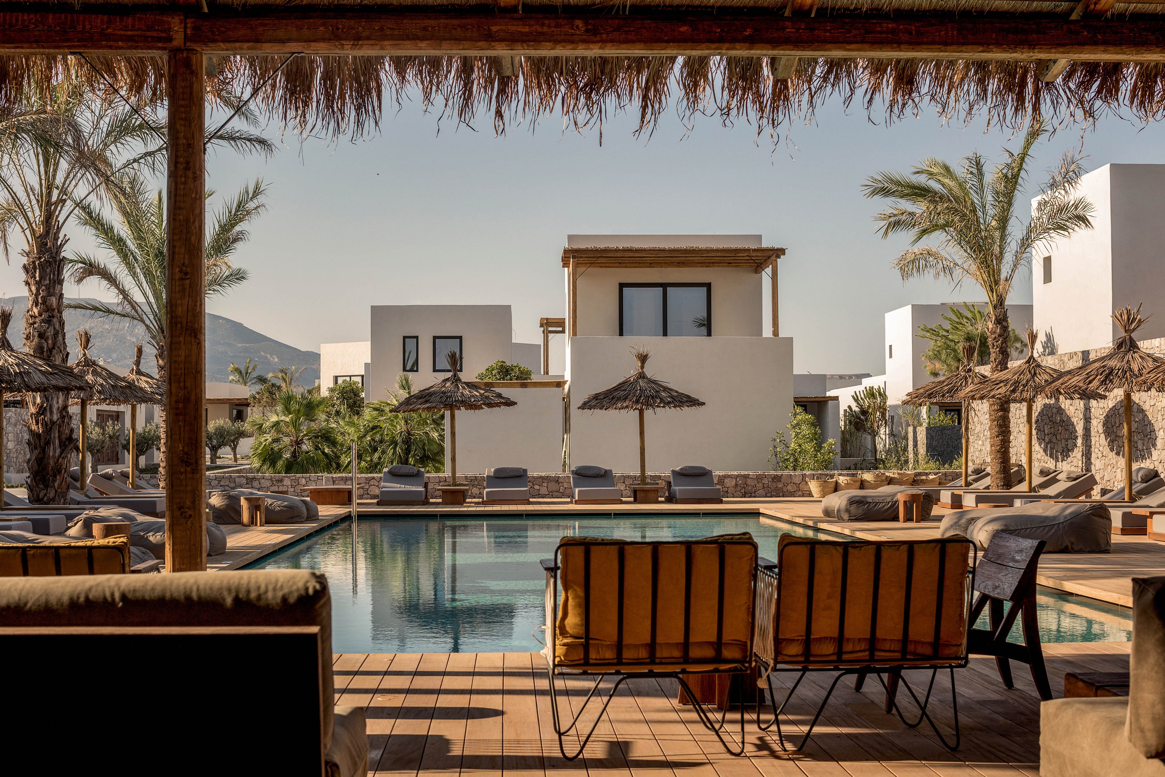 A contemporary resort pool area features cushioned lounge chairs and straw parasols under a shaded pergola. White minimalist buildings and palm trees surround the tranquil swimming pool, creating a relaxed vacation atmosphere. The scene is bathed in warm, natural sunlight with clear skies and distant mountains in the background.