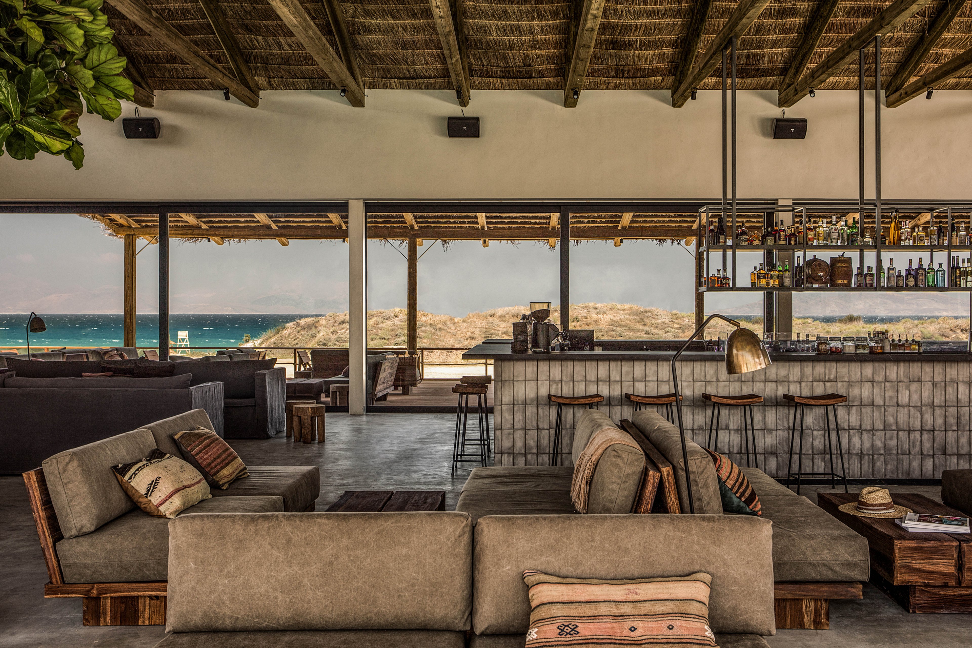 The image features a stylish lounge area with neutral-toned sofas and patterned pillows, set in front of a rustic bar. Large windows reveal a scenic ocean view and sandy dunes, enhancing the relaxed atmosphere. The space is characterized by natural wood elements, open ceilings, and soft lighting, creating a warm and inviting mood. No visible text or numeric values are present in the scene.